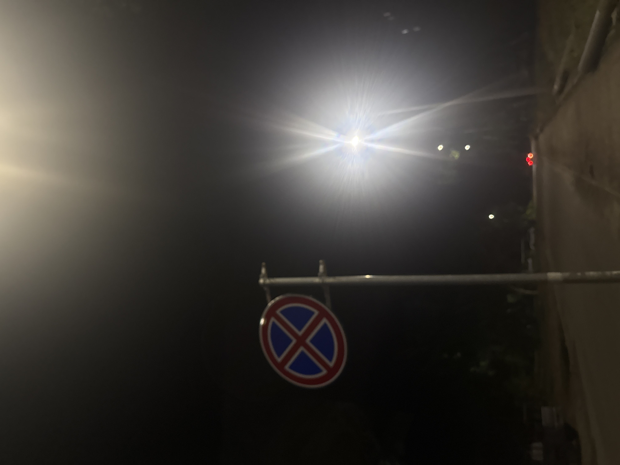 This is a night-time, low-angle, full-shot photograph of a street scene in Dnipro, Ukraine. The primary subject is a circular traffic sign, featuring a red border and a blue background with a red cross. This sign is mounted on a grey pole on the left side of the frame. To the right, a bright, out-of-focus street lamp with a lens flare dominates the upper right quadrant of the image. In the background, a dimly lit road stretches into the distance, with faint outlines of trees and buildings visible on either side. Two red taillights from a distant vehicle are visible on the road. The overall atmosphere is dark and somewhat hazy, likely due to the artificial lighting.