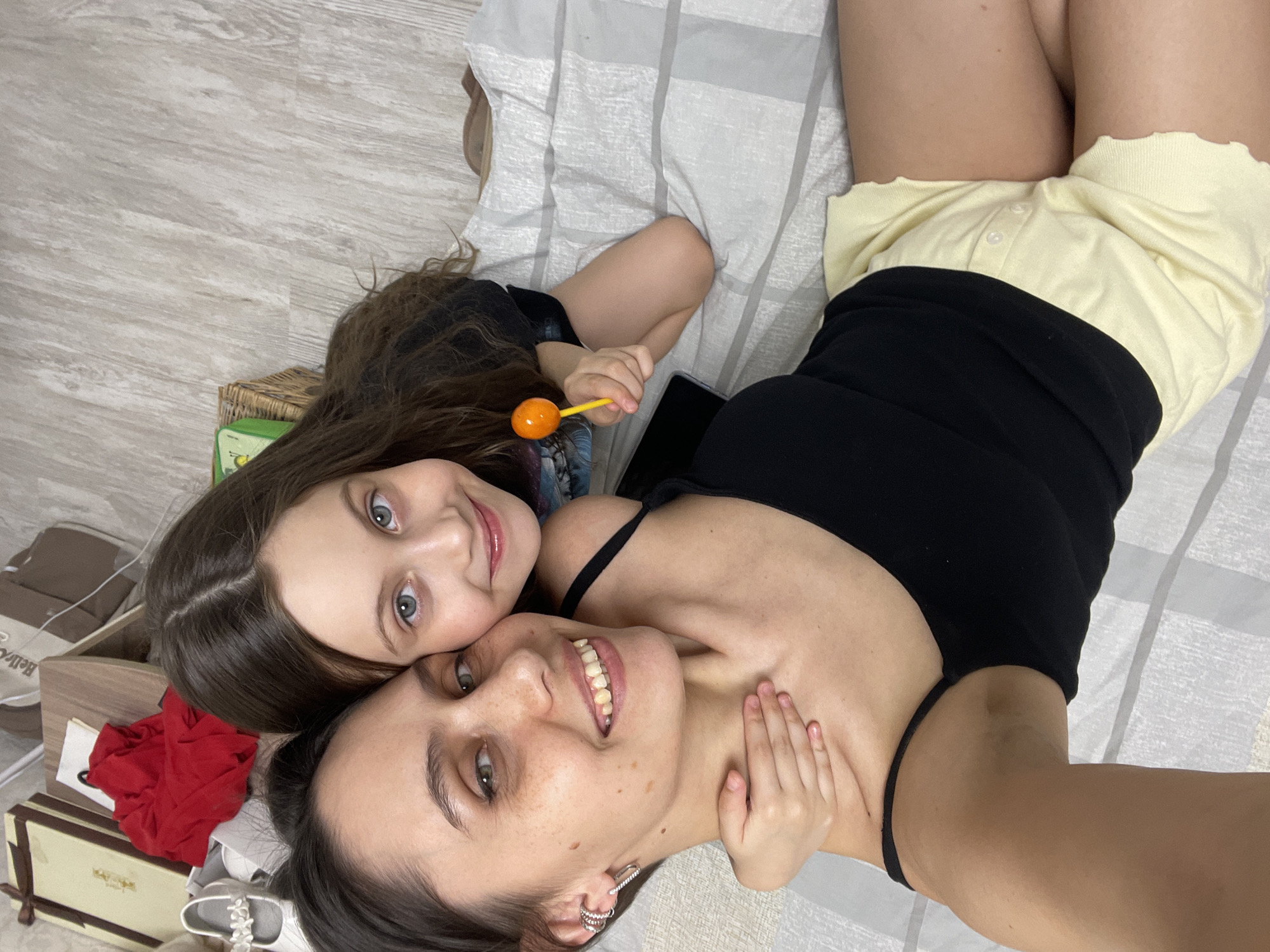 A selfie shows a woman and a young girl lying on a bed in a room. The woman is wearing a black tank top and yellow shorts, and the girl is wearing a black shirt. The girl is holding an orange lollipop. The bed has a grey and white striped sheet. The room has a wood floor and a side table with items on it including a red cloth, a pair of white shoes, and some boxes. Based on the user's information, the location is Izhevsk, Russia.