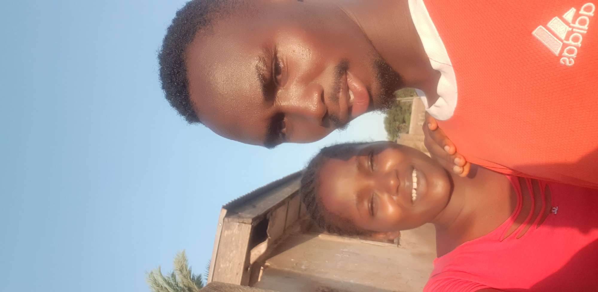 A close-up, brightly lit outdoor photograph depicts two dark-skinned individuals, a man and a woman, positioned for a selfie. The man, on the right, looks directly at the camera with a neutral expression. He has short, dark curly hair, a light beard, and is wearing an orange t-shirt with a mirrored "adidas" logo in white on the chest and a white trim collar. His right arm is extended, with his hand resting on the woman's left shoulder. The woman, positioned to the left and slightly behind the man, is smiling broadly with her eyes slightly squinted, likely due to the bright sunlight. She has dark hair styled in braids and is wearing a red top with three horizontal cut-outs on the chest and a small dark logo resembling the Chanel interlocking C. The background consists of a clear, bright blue sky dominating the upper half of the image. To the far left, a section of a palm tree frond is visible. Behind the woman, a tan-colored building with weathered wooden eaves and a section of corrugated metal roofing is partially seen. Distant green foliage is blurred in the mid-ground between the subjects. The location is Unknown, Unknown.