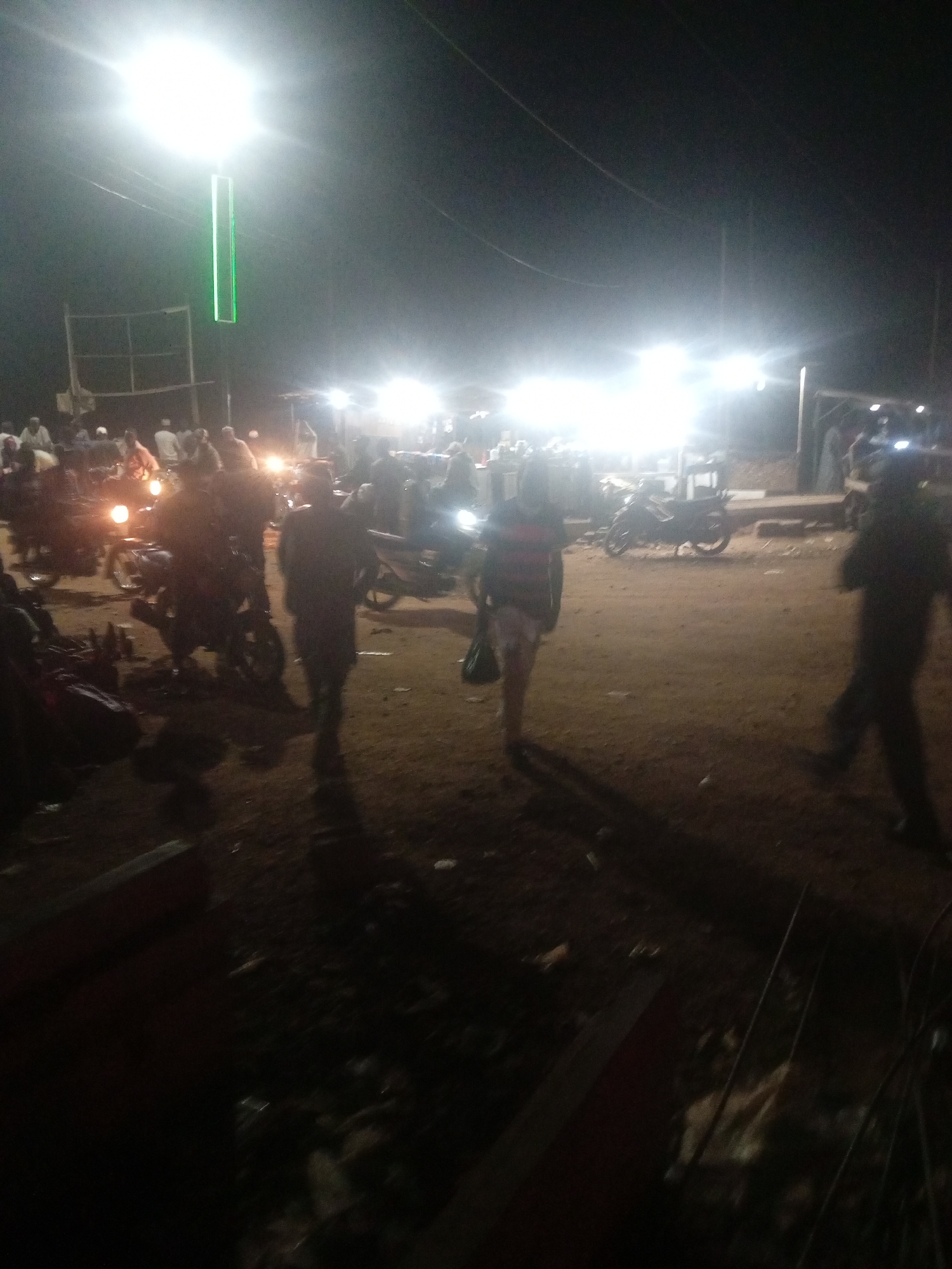 The image depicts an outdoor scene at night, likely in Tundun Wada West, Nigeria. The primary subjects are a group of people and several motorcycles, illuminated by bright artificial lights, suggesting a bustling night market or gathering. Several individuals are seen walking or standing amidst the crowd and motorcycles. One person in the foreground is walking away from the camera, while another, wearing a striped shirt, walks towards the camera. The overall atmosphere appears lively, with a sense of activity and movement, despite the low light conditions. The setting is an open, unpaved area, possibly a street or a market square, with some stalls or makeshift structures visible in the background. A tall, bright light fixture, possibly a street lamp, dominates the upper left portion of the image, casting a strong glare. A green illuminated vertical sign is also visible beneath the bright light. The weather is not discernible from the image. No visible text is present.