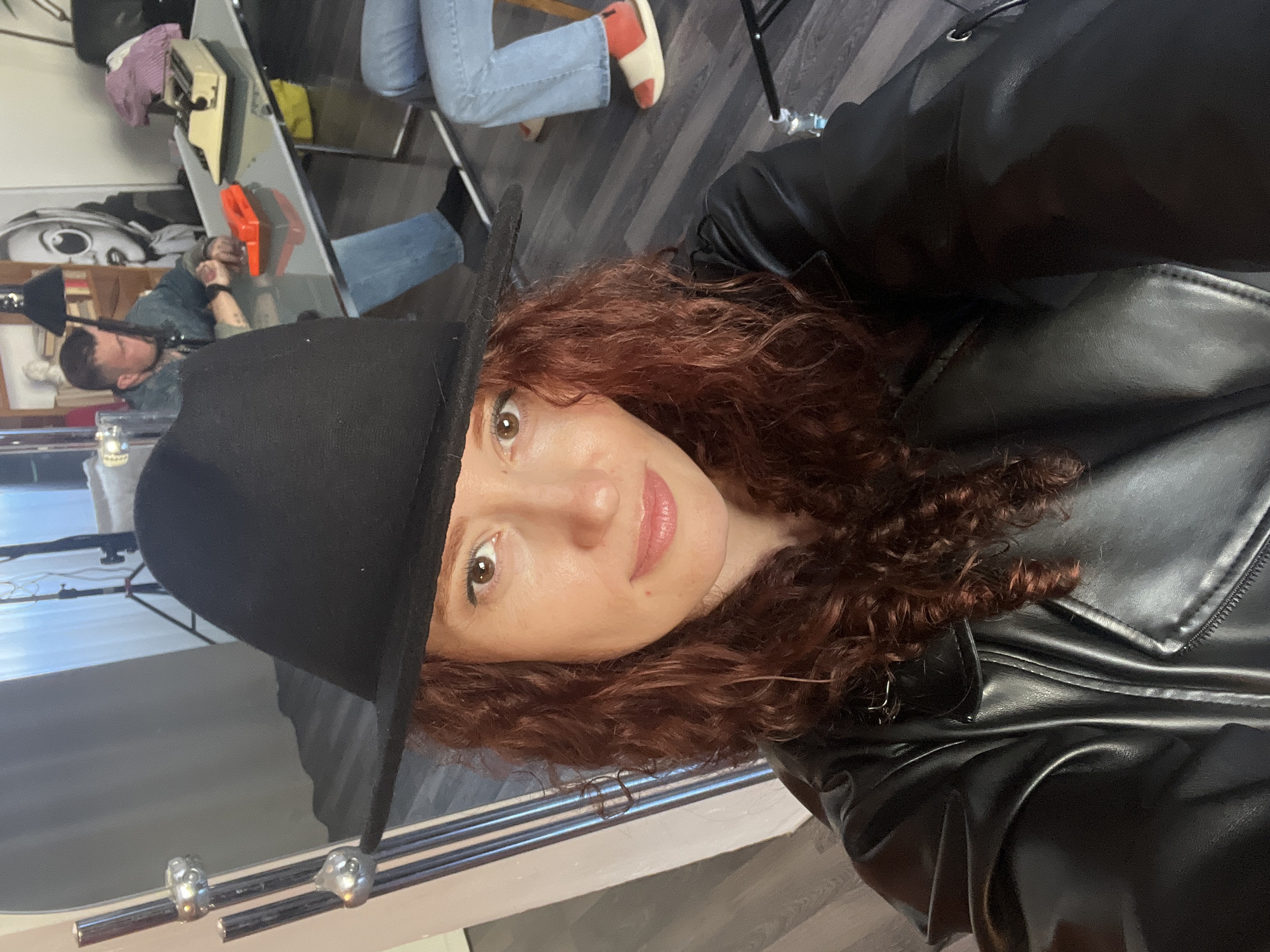 A woman with curly, auburn hair and fair skin is wearing a black hat and a black leather jacket. She is looking directly at the camera. In the background, there is a man lying on a table with various objects around him, including what appears to be a typewriter. Other people are sitting around. The image may have been taken indoors. It is not possible to determine the location in Saint Petersburg, Russia, or any specific actions or interactions from this single image.