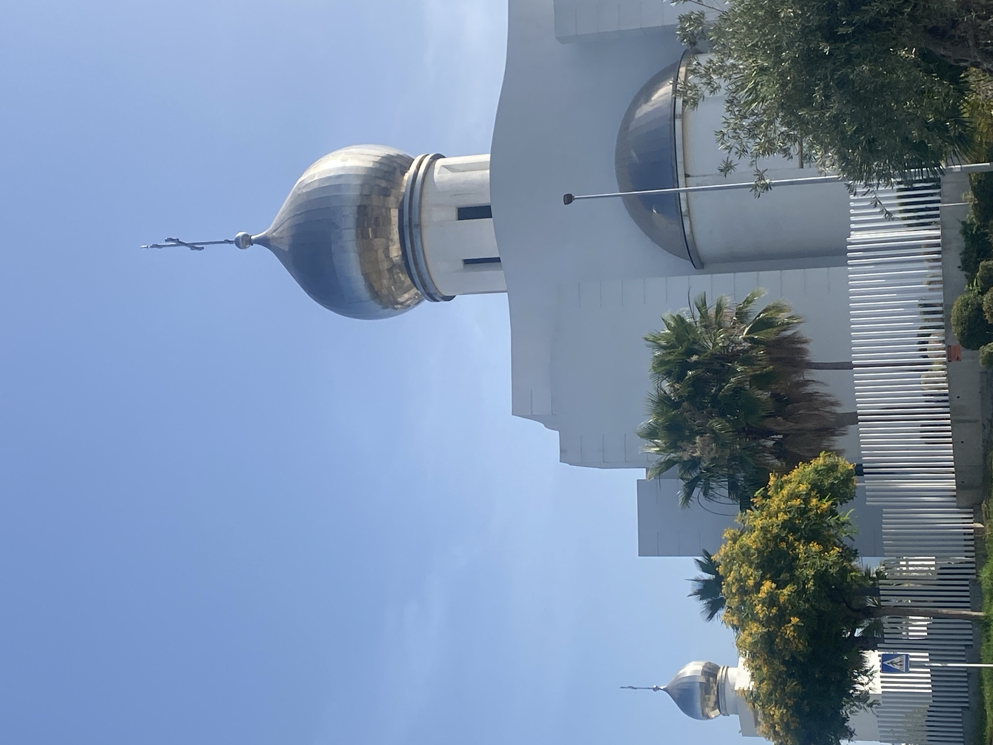 The image shows the Russian Orthodox Church of the Archangel Michael in Estepona, Spain, under a clear blue sky. The prominent feature is the church's large, silver-colored dome topped with a cross. The building is modern and white, with a distinct architectural style. In the foreground, there are several palm trees and a tree with yellow blossoms, behind a white picket fence. To the right of the main dome, another smaller dome is visible, partially obscured by trees and a street lamp. The overall scene depicts a sunny day in a location with Mediterranean vegetation.