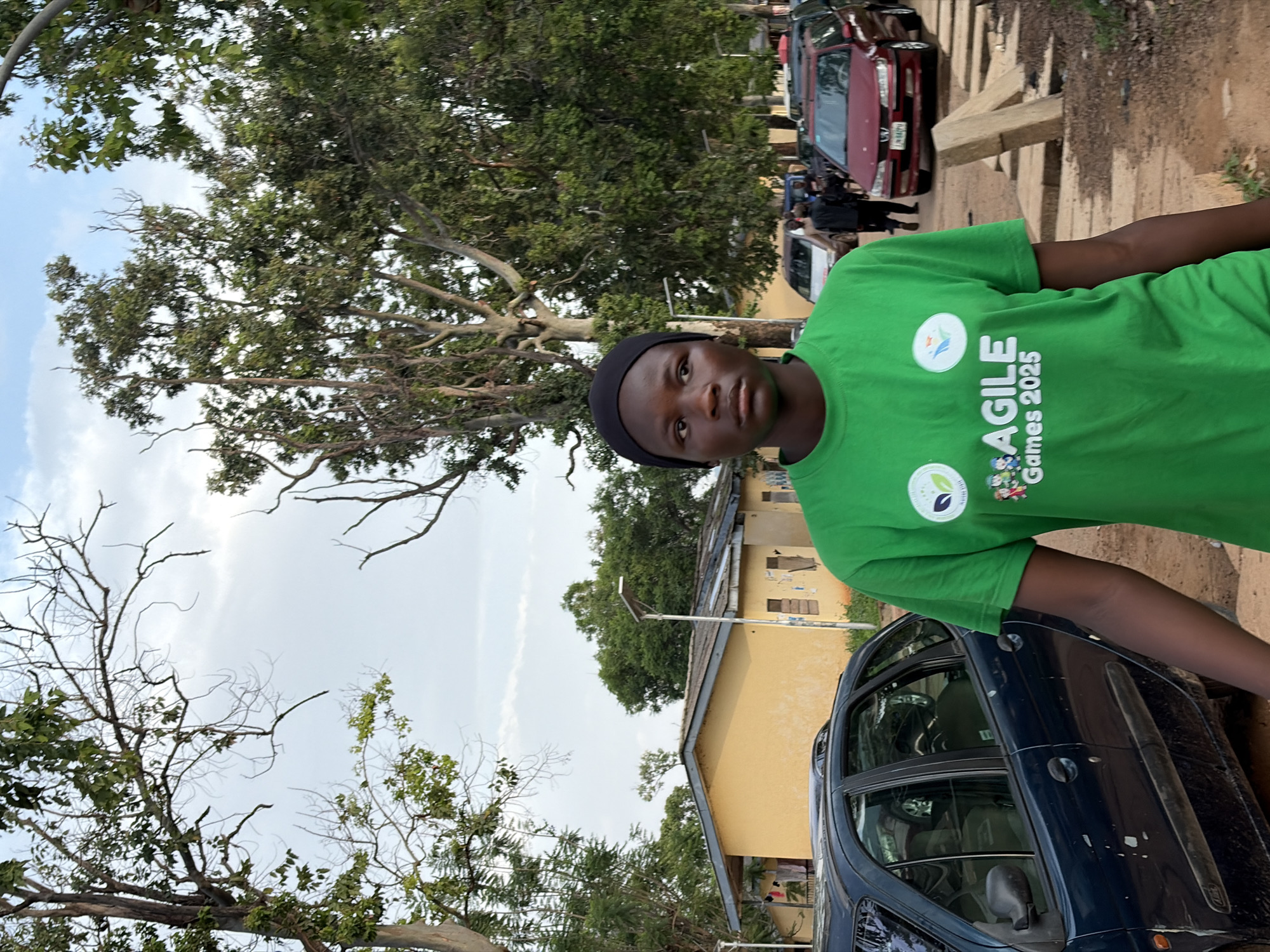 A young person wearing a bright green t-shirt with the text "AGILE Games 2025" printed on it stands in the foreground, looking to the left. The t-shirt also features two circular emblems. The person is wearing a black beanie. Behind them, a yellow building with a grey roof is visible, along with trees and a cloudy sky. Several cars are parked in the background, including a dark blue car in the left foreground and a maroon car to the right. The scene appears to be outdoors in a somewhat rural or suburban area, possibly in Rafin Par, Nigeria, given the architecture and vegetation. The ground is a dirt path with some stone elements.
