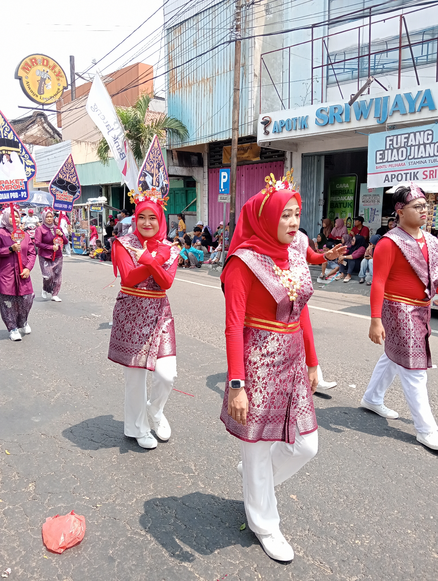 An outdoor, eye-level, medium shot captures a parade in progress on a street in Demak, Indonesia. Three women in red headscarves and traditional outfits, adorned with gold and intricate red patterns, are walking from left to right. The woman in the foreground, wearing a red top, a patterned skirt, and white pants, is the most prominent. She is smiling and gesturing with her hands. Behind her, two other women, dressed similarly in burgundy, are also walking. A man in the background, also in a traditional outfit and headwear, is walking to the right. The street is paved, with cracks visible. Shops line the street, one with a prominent blue sign reading "APOTIK SRIWIJAYA". Spectators are seated and standing on the sidewalk, watching the parade. The sky is bright and clear. A crumpled red plastic bag lies on the street in the foreground.