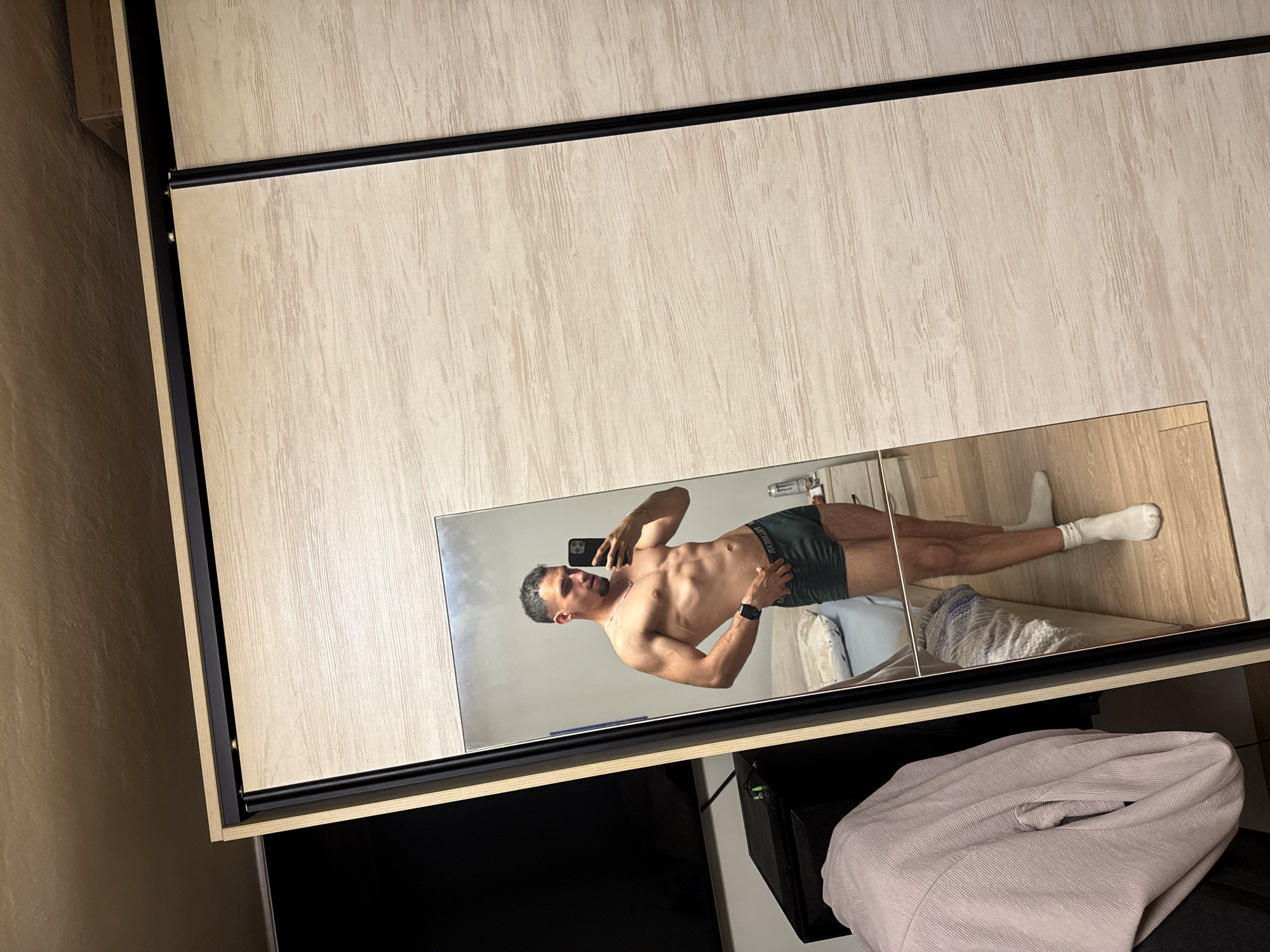 A man wearing socks and dark boxer briefs stands in front of a mirror and takes a photo of himself. The mirror is part of a wardrobe, which is placed in a room. A television and a pile of clothes sit beneath the wardrobe. The location is likely in a residence in Shumen, Bulgaria.