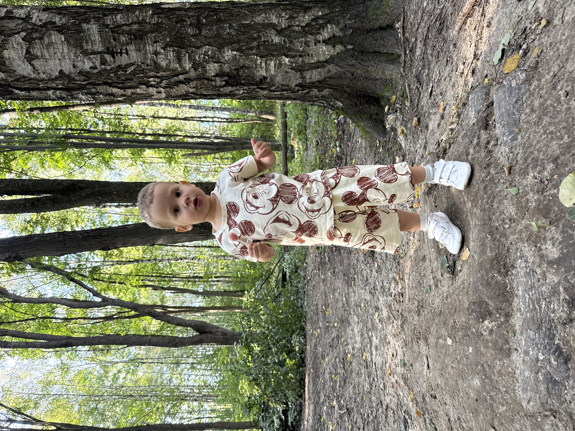 A young child, appearing to be between two and three years old, stands on a dirt path in a wooded area. The child has light brown hair and is wearing a light-colored t-shirt and shorts with a brown pattern of circular shapes. The child's feet are clad in white velcro sneakers and grey socks. The child holds a pink lollipop in their right hand and their left hand is extended slightly. Behind the child, the ground is covered in dirt, leaves, and small stones. Several trees with green foliage and grey bark are visible in the background, suggesting a natural, outdoor environment. The lighting indicates it is daytime, with sunlight filtering through the trees. The scene is consistent with a park or forest area within or on the outskirts of the city of Dumino, Russia.