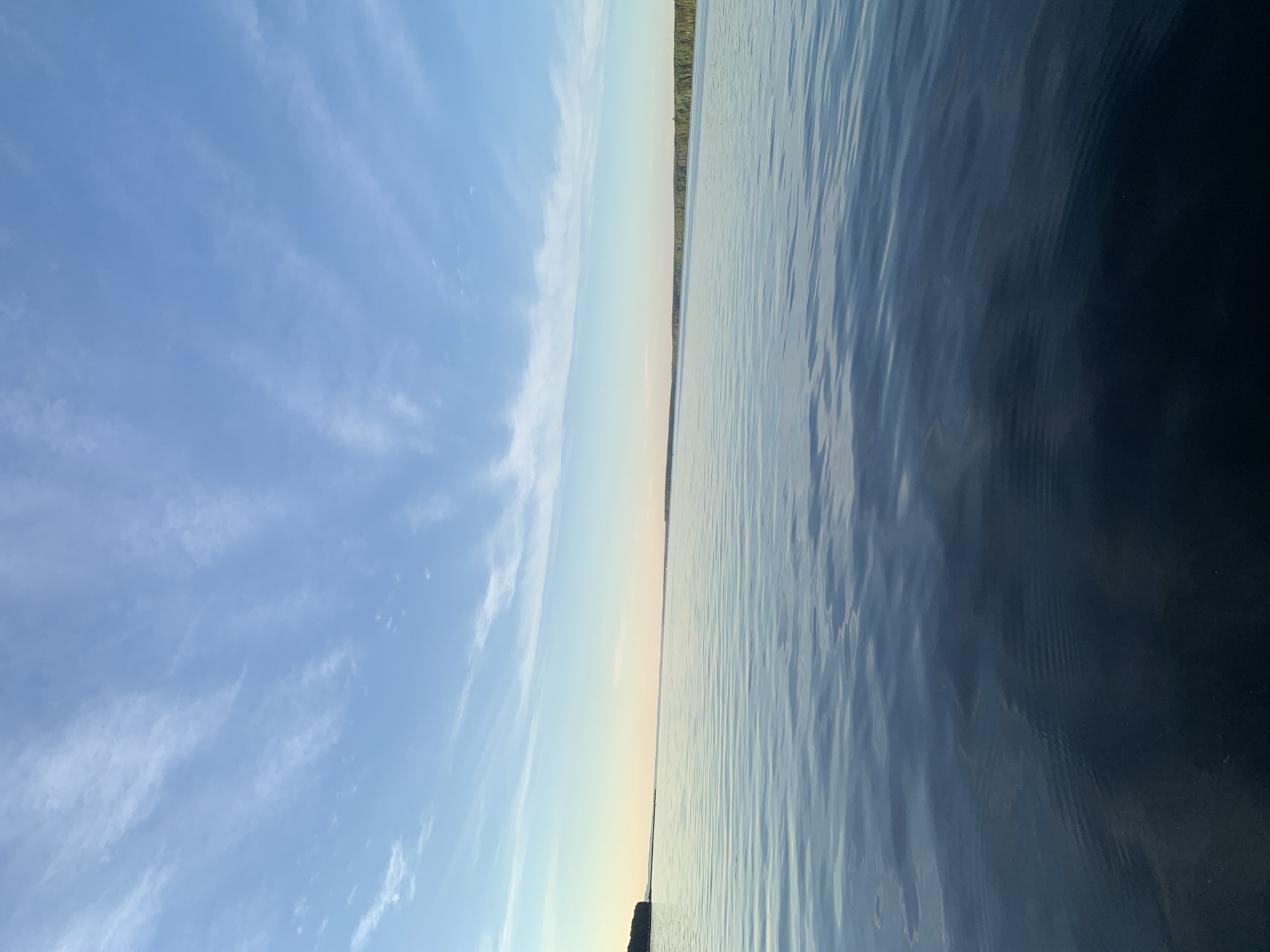 An outdoor, vertical, eye-level, full shot captures a wide view of the Kama River at sunset in Chaikovsky, Russia. The sky above is a pale blue, transitioning to a soft orange hue near the horizon. Wispy white clouds stretch across the sky, catching the last rays of the sun. The surface of the river is calm, with gentle ripples reflecting the colors of the sky. In the distance, the far bank of the river is lined with a dense forest, dark green and silhouetted against the fading light. A small section of land is visible on the left, also covered in trees. The water in the foreground is darker, appearing deep and expansive. There are no people or man-made structures visible in this natural landscape.