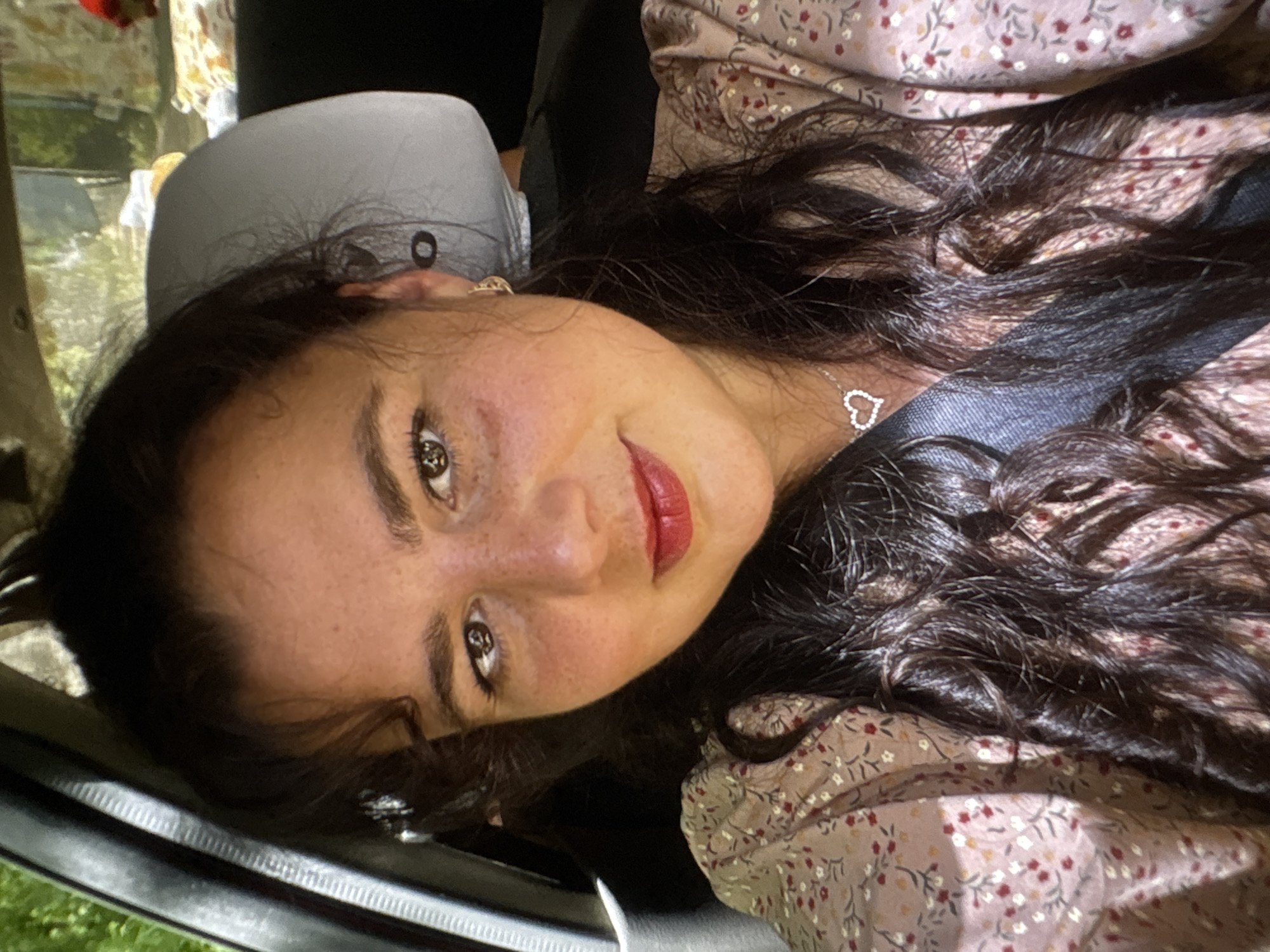 This is a close-up, sideways photograph of a young woman. She has long, dark, wavy hair, fair skin with freckles, and brown eyes. She is wearing a light-colored, floral-patterned top and a silver necklace with a heart pendant. Her lips are painted red, and she is smiling slightly. The background appears to be inside a vehicle, with a gray headrest visible behind her. The lighting suggests it is daytime, but the specific location and weather are not discernible. The image conveys a sense of casualness and contentment. There is no visible text in the image.