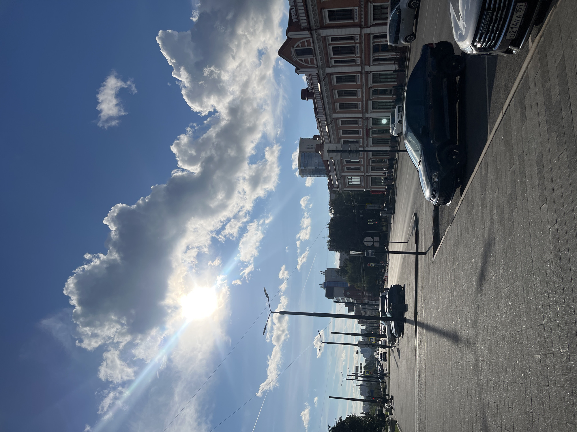 The image shows a city street in Perm, Russia, on a sunny day. The sky is blue with fluffy white clouds, and the sun is shining brightly, casting long shadows. The street is lined with buildings, some of which appear to be historic with ornate architecture, while others are modern high-rises. Cars are visible on the road, including a black sedan and a white SUV. The sidewalk is paved with dark gray tiles, and there are several streetlights and traffic signs. In the distance, a few people can be seen walking on the sidewalk and crossing the street. The overall impression is of a bustling urban environment on a clear day. The text on the building in the upper right corner appears to be in Cyrillic.