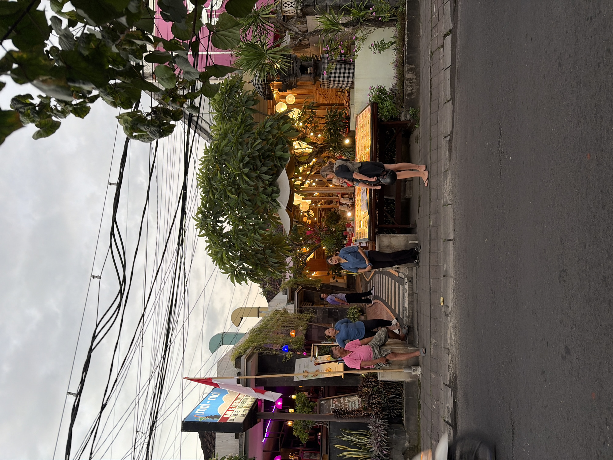 In Kuta Selatan, Indonesia, a street scene unfolds in front of a bar and restaurant. The establishment, named "Mai-Mai," features a vibrant facade with a prominent sign displaying the restaurant's name and "Bar & Restaurant." A Indonesian flag, red and white, flutters on a pole outside. The building is adorned with hanging plants and illuminated by numerous globe lights, creating a warm ambiance. A wooden table displays menus, and various people are visible.
On the left, a man in a pink t-shirt and checkered shorts sits on a low wall. Beside him, a woman in a blue collared shirt and dark pants stands. Further back, a man in a purple shirt stands near the entrance of the establishment. On the right side of the image, a woman in a black dress holds a child, and another woman in dark pants and a blue top stands near a table laden with what appear to be food items. A dark-colored motorcycle is captured in motion on the left side of the asphalt road, indicating traffic. Overhead, a dense network of black electrical wires crisscrosses the sky, partially obscuring the overcast sky. The sidewalk is paved with light-colored rectangular stones, and a border of low-lying plants runs along the front of the establishment. A black and white checkered fabric is draped over a stone structure with a peaked roof, suggesting a traditional Balinese architectural element. The overall scene captures a moment in the evening in this bustling Indonesian city.
