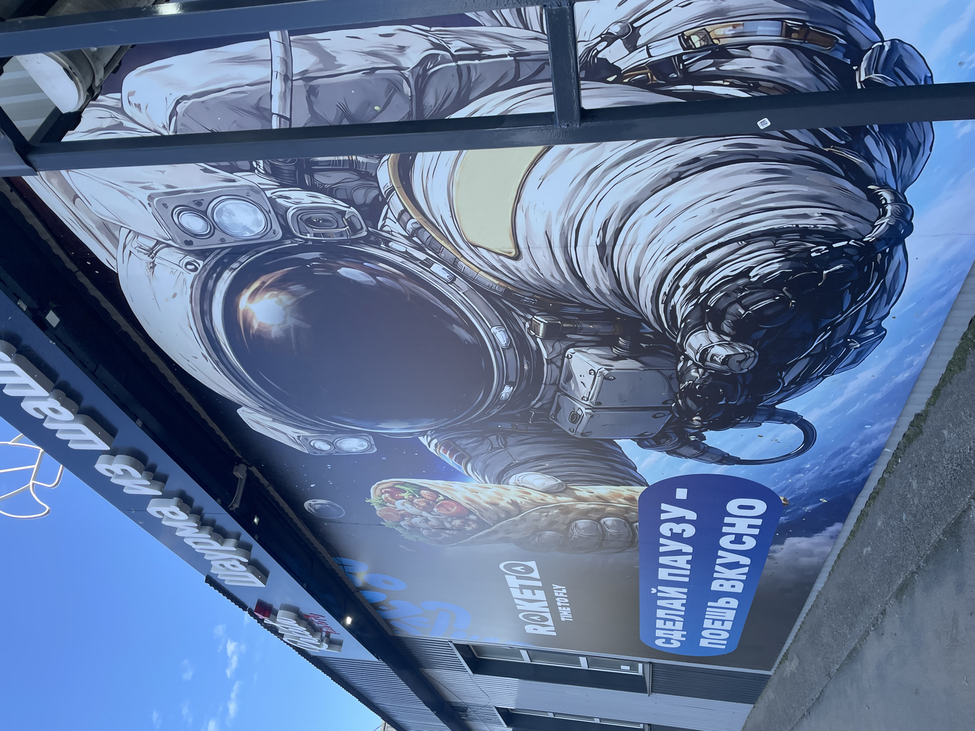 The image shows a building in the city of Chaikovsky, Russia, with a large mural on its exterior. The mural depicts an astronaut in a spacesuit holding a large shawarma. Above the mural, in white lettering, is the text "Шаурма из Шау" which translates to "Shawarma from Shau." Below the astronaut, on a blue background, are the words "СДЕЛАЙ ПАУЗУ - ПОЕШЬ ВКУСНО" which translates to "Take a break - eat deliciously." To the left of the astronaut is a logo that reads "ROKETO TIME TO FLY." The building appears to be a commercial establishment, possibly a fast-food restaurant, given the imagery and text. The sky above is blue with some clouds.