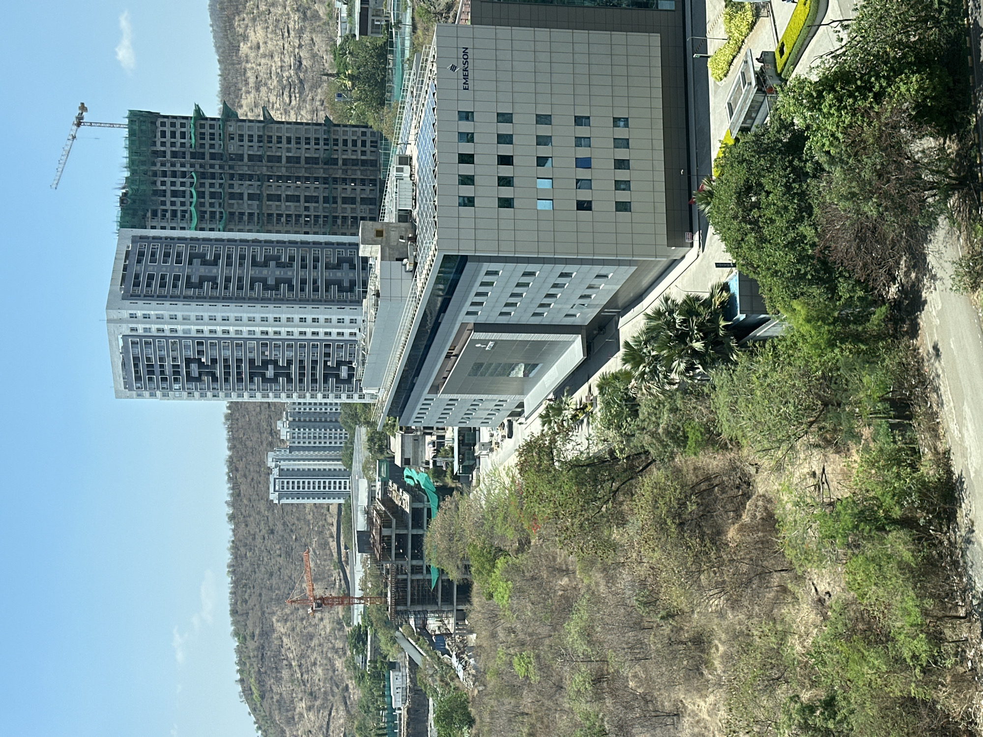 The image shows a cityscape in Marunji, India, with a mix of developed and developing areas. Multiple high-rise buildings are visible, some under construction with cranes present. "EMERSON" is prominently displayed on one of the completed buildings. The area also features a hillside, trees, and a road. The sky is clear and blue.