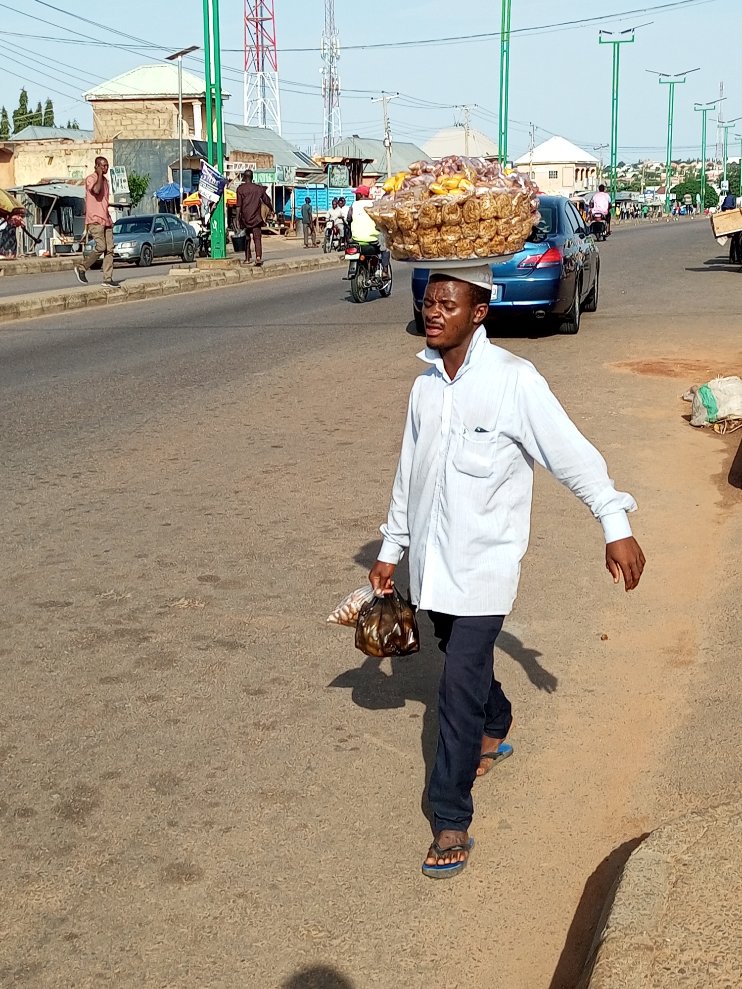A man walks down a paved road in Tempure, Nigeria, carrying a large platter of packaged goods on his head and a plastic bag of goods in his hand. He is wearing a light blue collared shirt and dark pants. The road is lined with buildings and other pedestrians. A blue car is parked on the right side of the road. The sky is clear and the sun is shining.