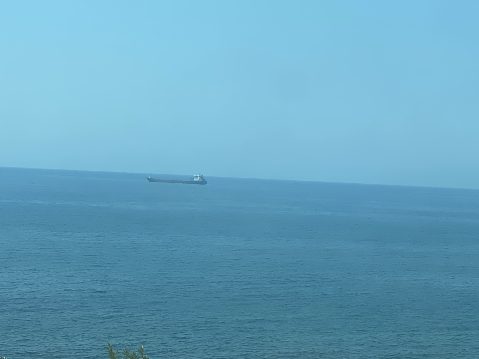 A cargo ship is visible in the distance on a calm, blue sea under a clear, light blue sky. The ship is positioned horizontally in the frame, with its bow pointing to the left. The vast expanse of the sea dominates the lower half of the image, while the sky occupies the upper portion. The scene suggests an open water environment, likely near the coast of Bar, Montenegro, given the characteristic color of the Adriatic Sea and the clear sky often seen in the region. There are no people or other objects clearly discernible in the image, emphasizing the solitary nature of the ship.