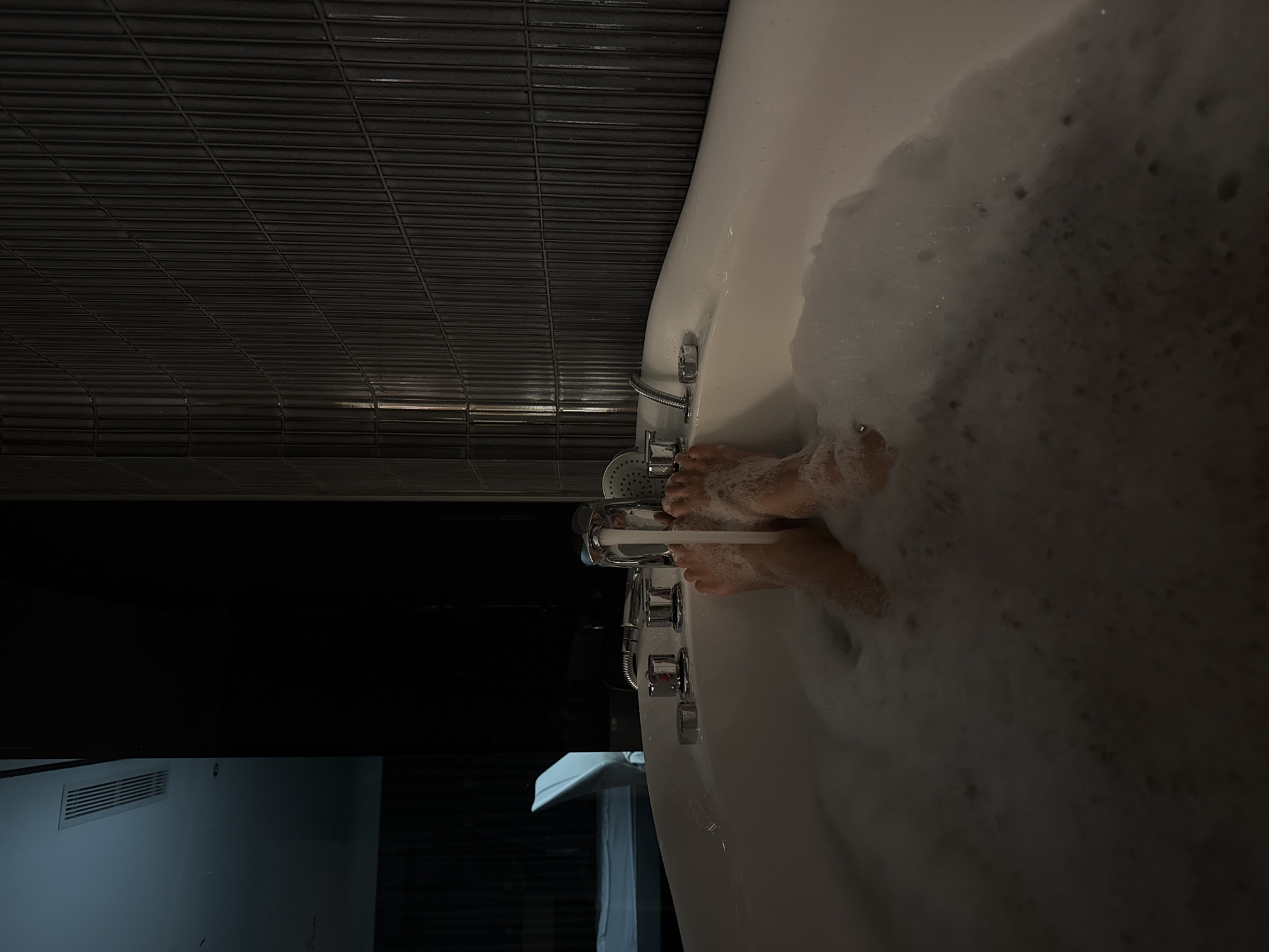 Here is a detailed description of the image:
A sideways view of a bubble bath with a person's feet visible among the suds. The tub is white and appears to be a modern design. The bathtub faucet is on, and water is running into the tub. The bathroom wall is covered in dark-colored, horizontal tiles. A blurry image of the bathroom can be seen in the background, featuring a bright wall and an air conditioning unit.