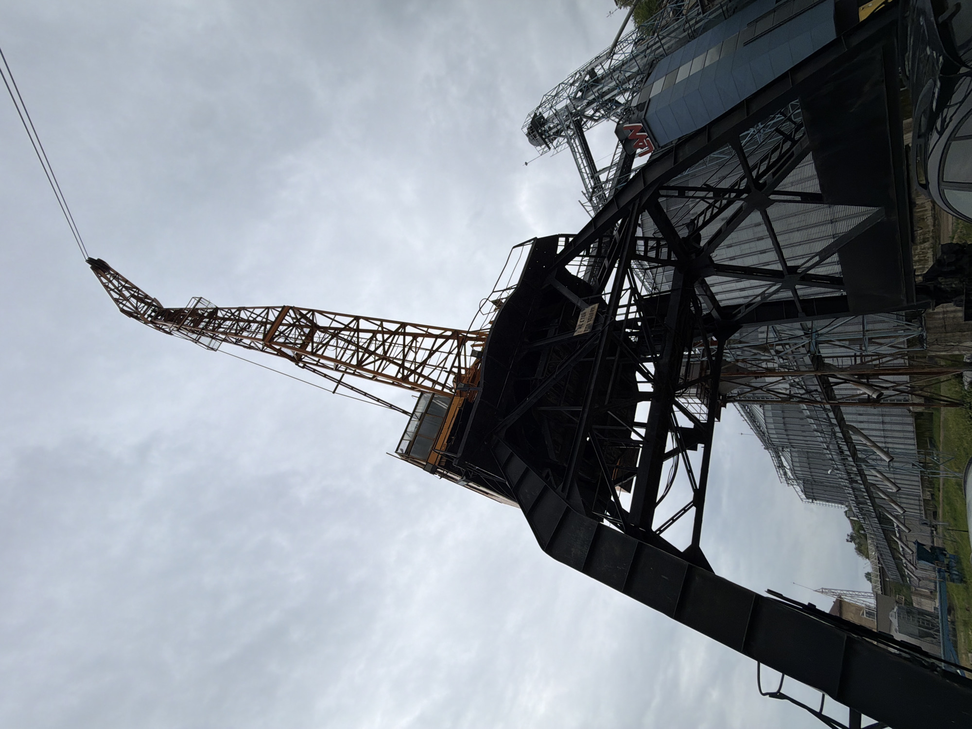 An angled low-angle shot depicts a crane situated at the Port of Svishtov in Bulgaria. The crane, with its lattice boom extending to the left, exhibits a combination of rust-orange on the boom's skeletal structure and black on the base and supporting framework. Adjacent to the crane, a complex of metallic structures, possibly grain silos, rises against a cloudy sky. The ground below features green vegetation and hints of industrial buildings in the distance.