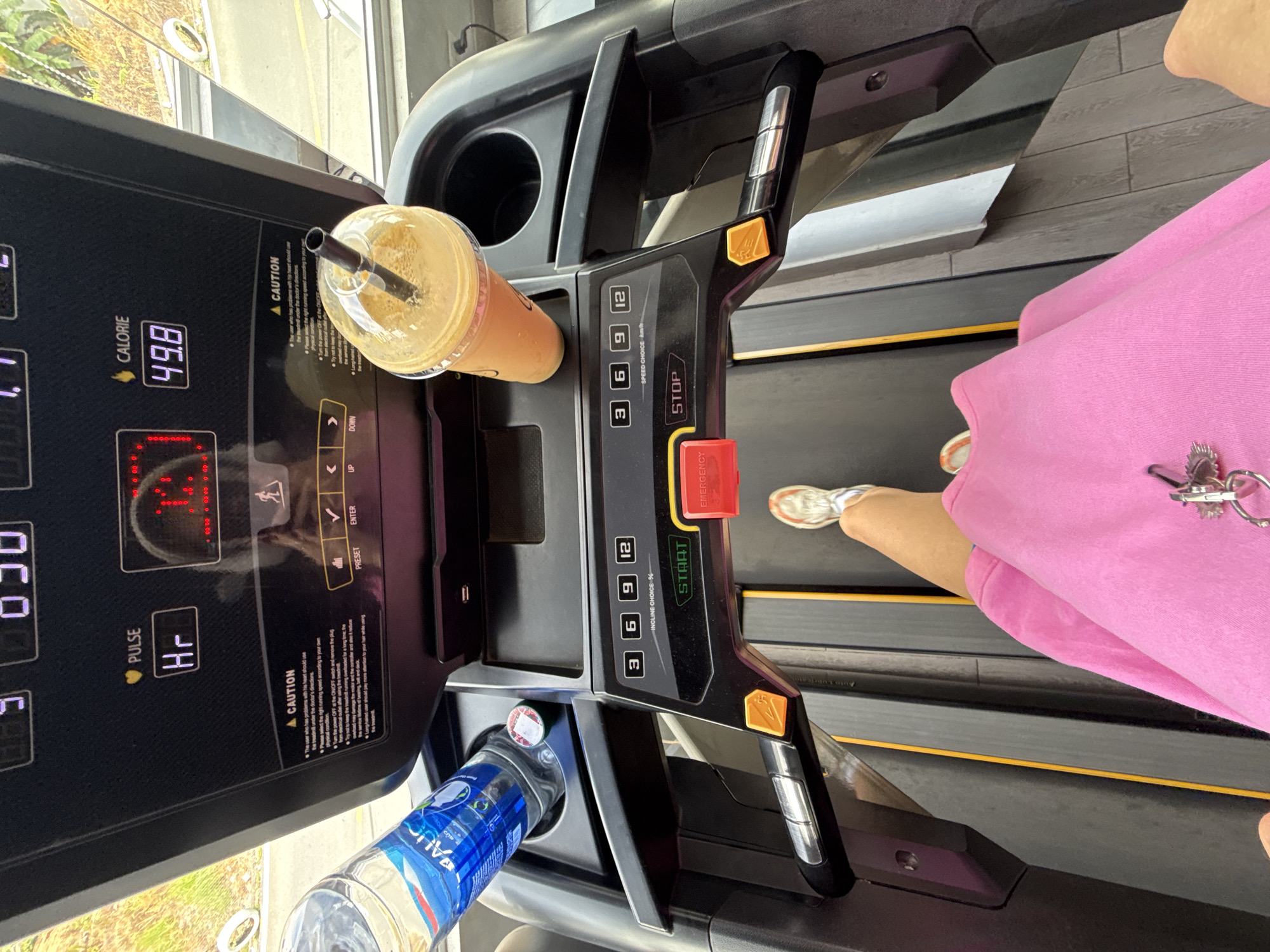 The image shows a first-person perspective of a person on a treadmill in a gym. The person is wearing white sneakers and a bright pink outfit. They are holding keys in their hand. On the treadmill console, a digital display shows "49.8" calories burned. To the left of the console is a plastic bottle of water. To the right is a plastic cup with a beverage and a straw. The background appears to be a window looking out onto a natural outdoor environment with trees and sunlight. The floor of the gym is made of dark wood planks. The image is taken in Da Lat, Vietnam.