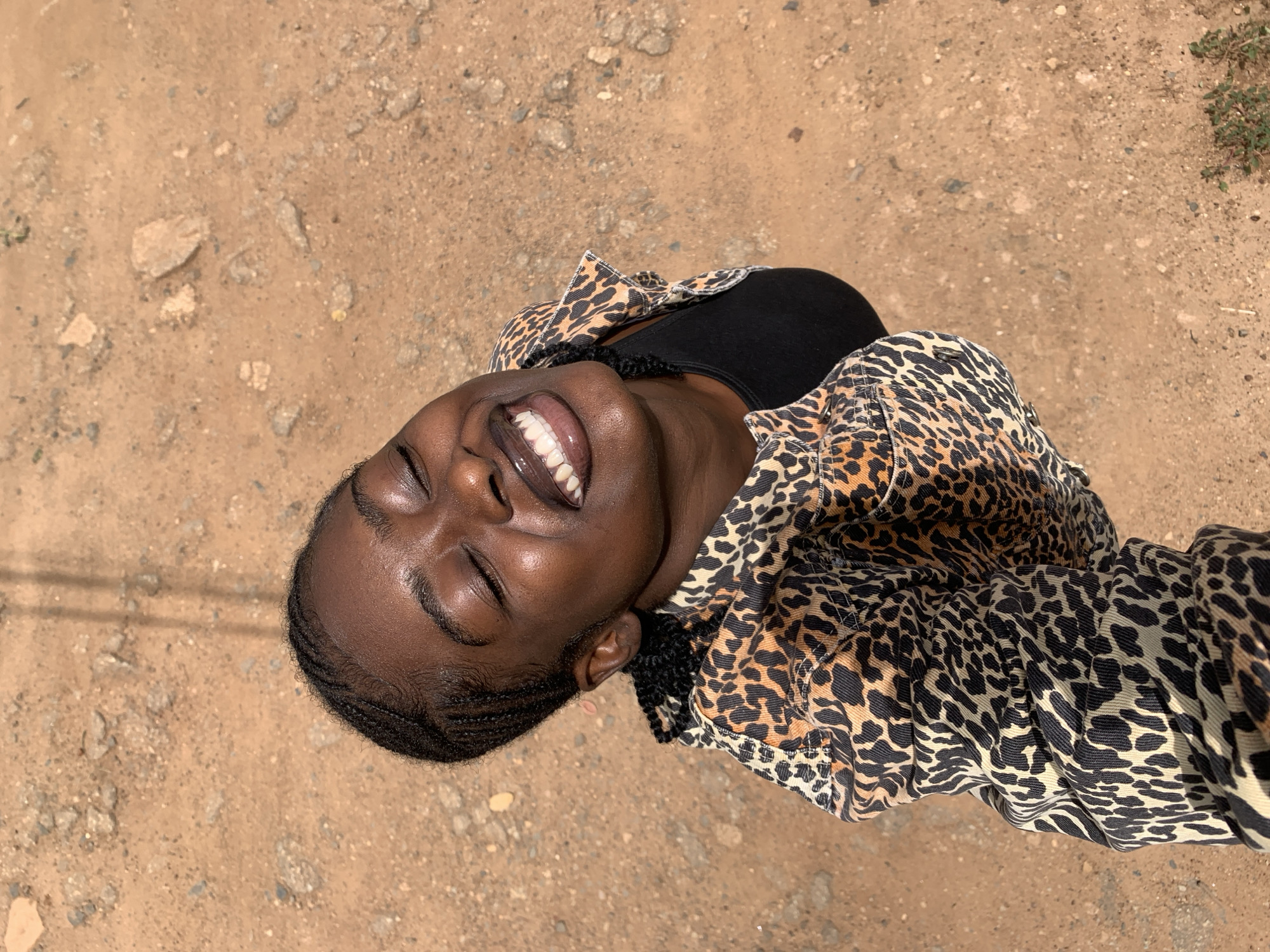 A dark-skinned female subject is depicted from an elevated perspective, positioned centrally and looking upwards with closed eyes and a broad smile, revealing white teeth and glossy lips. Her hair is styled in tight, dark braids. She wears an open, long-sleeved jacket with a black and brown leopard print pattern over a plain black top. The background is an unpaved, light brown sandy or dirt ground interspersed with numerous small, grey and brown rocks and pebbles. Faint vertical shadows are visible on the ground in the upper half of the image, and minimal green vegetation is present in the lower right corner. The scene is outdoors under bright sunlight in Rafin Par, Nigeria.