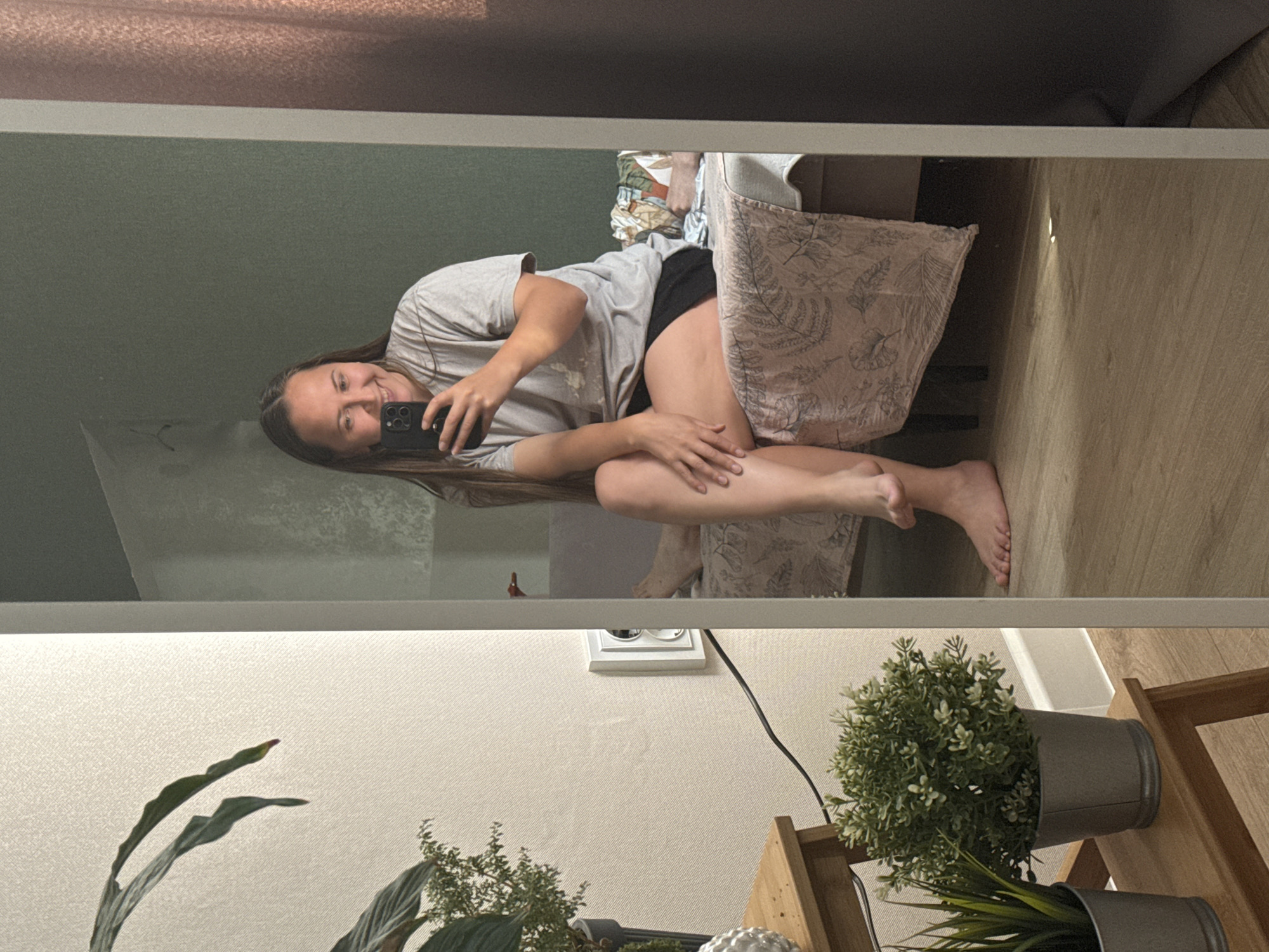 A woman is taking a selfie in a mirror. She is sitting on a chair in what appears to be a room. The background shows a wall with an electrical outlet and a shelf with potted plants. There is no information to indicate this photo was taken in Izhevsk, Russia.