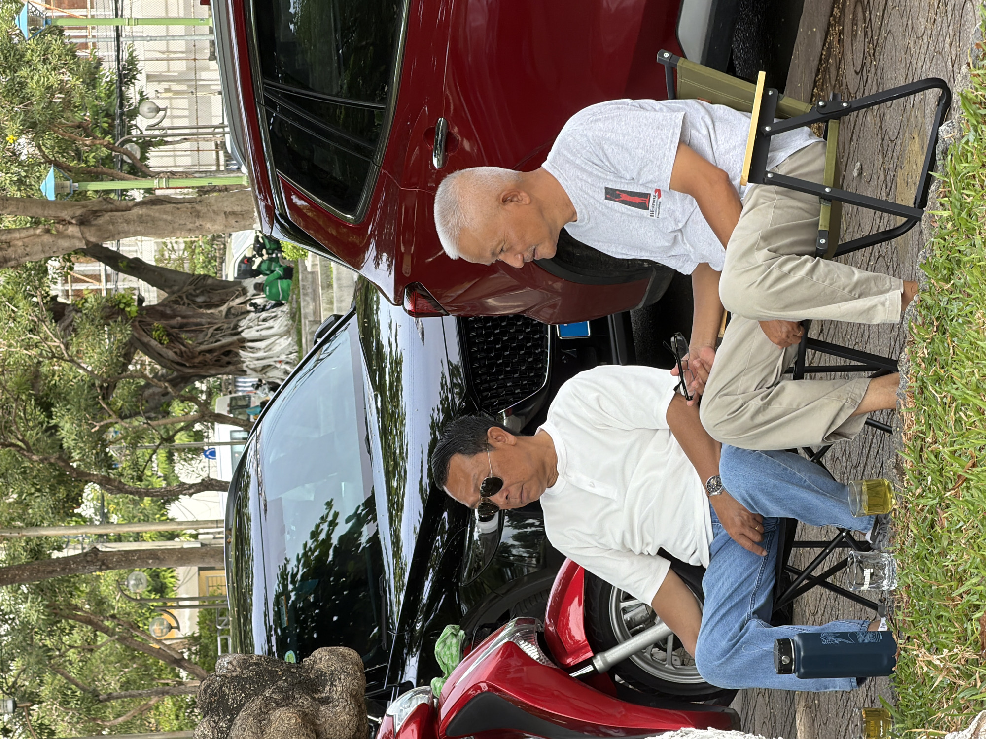 Two middle-aged Asian men are seated on foldable chairs in an outdoor urban setting. The man on the left wears sunglasses, a white polo shirt, and blue jeans. He is holding a pair of dark-rimmed glasses. The man on the right has short grey hair and wears a light grey t-shirt with a graphic and tan pants. He is looking to his left. In front of them are two glasses of a golden liquid and a dark blue water bottle. Behind them, a red SUV and a black SUV are parked, with the front of a red motorcycle visible on the left. The background consists of trees and buildings. This scene is likely in Nha Trang, Vietnam, given the style of the cars, motorcycle, clothing, and the presence of mature trees commonly found in the city's parks or along its streets.