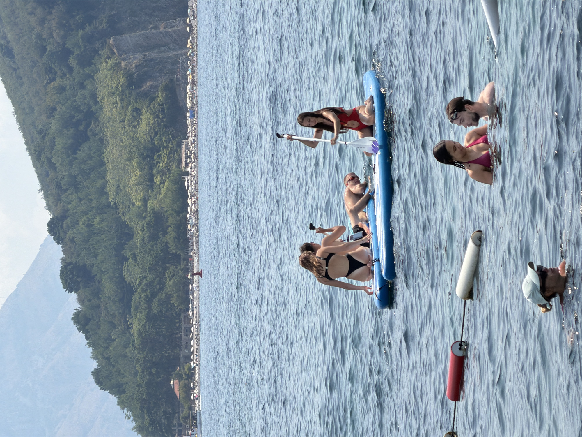 The image depicts a sunny day at a beach in Budva, Montenegro. In the foreground, two people are partially submerged in the clear blue water, looking at each other. Further out in the sea, a group of people are on two inflatable paddleboards. On one paddleboard, three individuals are seated or lying down, while another person is standing and holding a paddle. On the second paddleboard, a person in a red swimsuit is kneeling and holding a paddle. In the background, a beach lined with umbrellas and lounge chairs can be seen, with lush green hills and mountains rising behind it. The overall scene conveys a relaxed and enjoyable summer atmosphere.