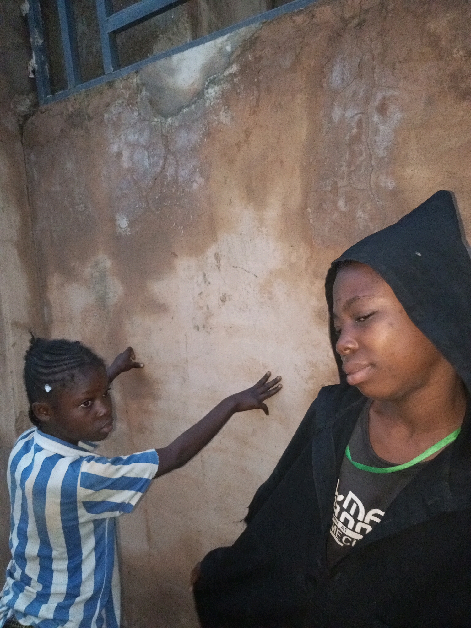 In Tempure, Nigeria, two young individuals stand against a textured, light brown wall. The individual on the left, a child with braided hair, wears a blue and white striped shirt and extends their left arm, hand open. To the right, another young person is partially visible, wearing a black hooded garment over a dark t-shirt with white lettering. A thin, green necklace is visible around their neck. Above them, a section of a dark blue metal railing is present, suggesting an outdoor setting possibly adjacent to a building. The wall's surface displays patches of lighter discoloration and faint cracks.