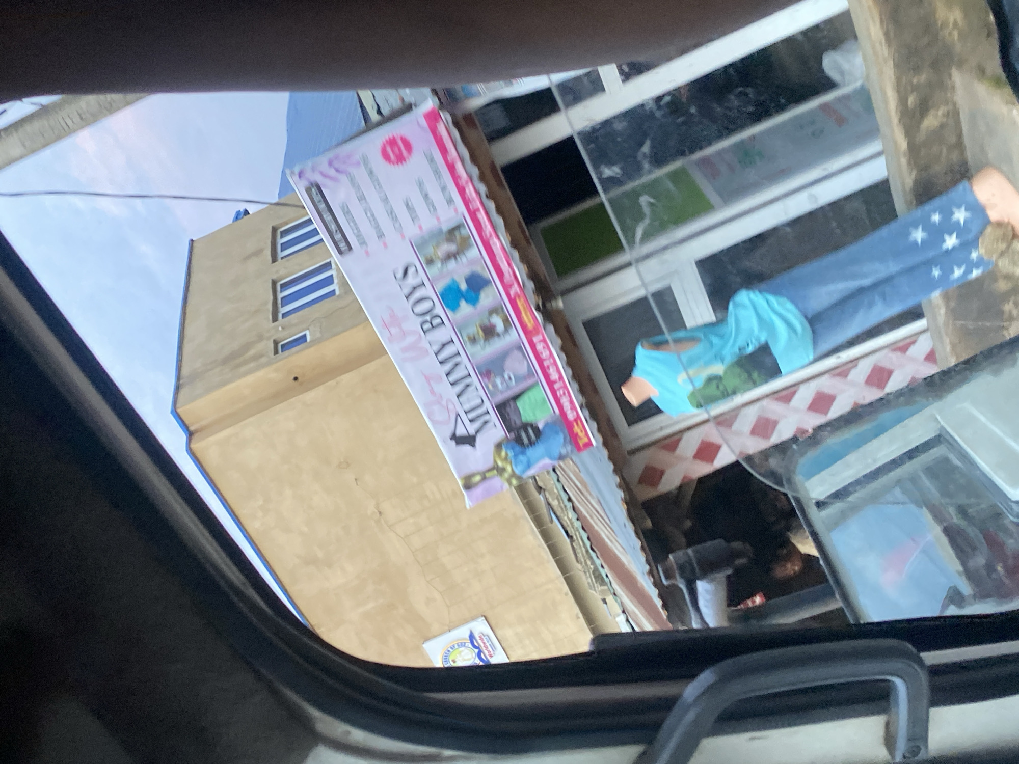 The image depicts a street scene in Ikorodu, Nigeria, captured from inside a vehicle. A prominent sign for "MUMMY BOYS" is visible, advertising children's clothing with images of outfits and a phone number. Below the sign, a stall with a corrugated iron roof displays red and white checkered patterns. A mannequin dressed in a blue sleeveless shirt and star-patterned pants stands outside the stall, positioned near a window with green and white panes. In the background, a tan building with multiple windows stands against a slightly overcast sky. A smaller sign for "CHURCH OF GOD WORLDWIDE" is affixed to the building. The lower portion of the image shows parts of the vehicle interior, including a black handle and a reflection on the window.