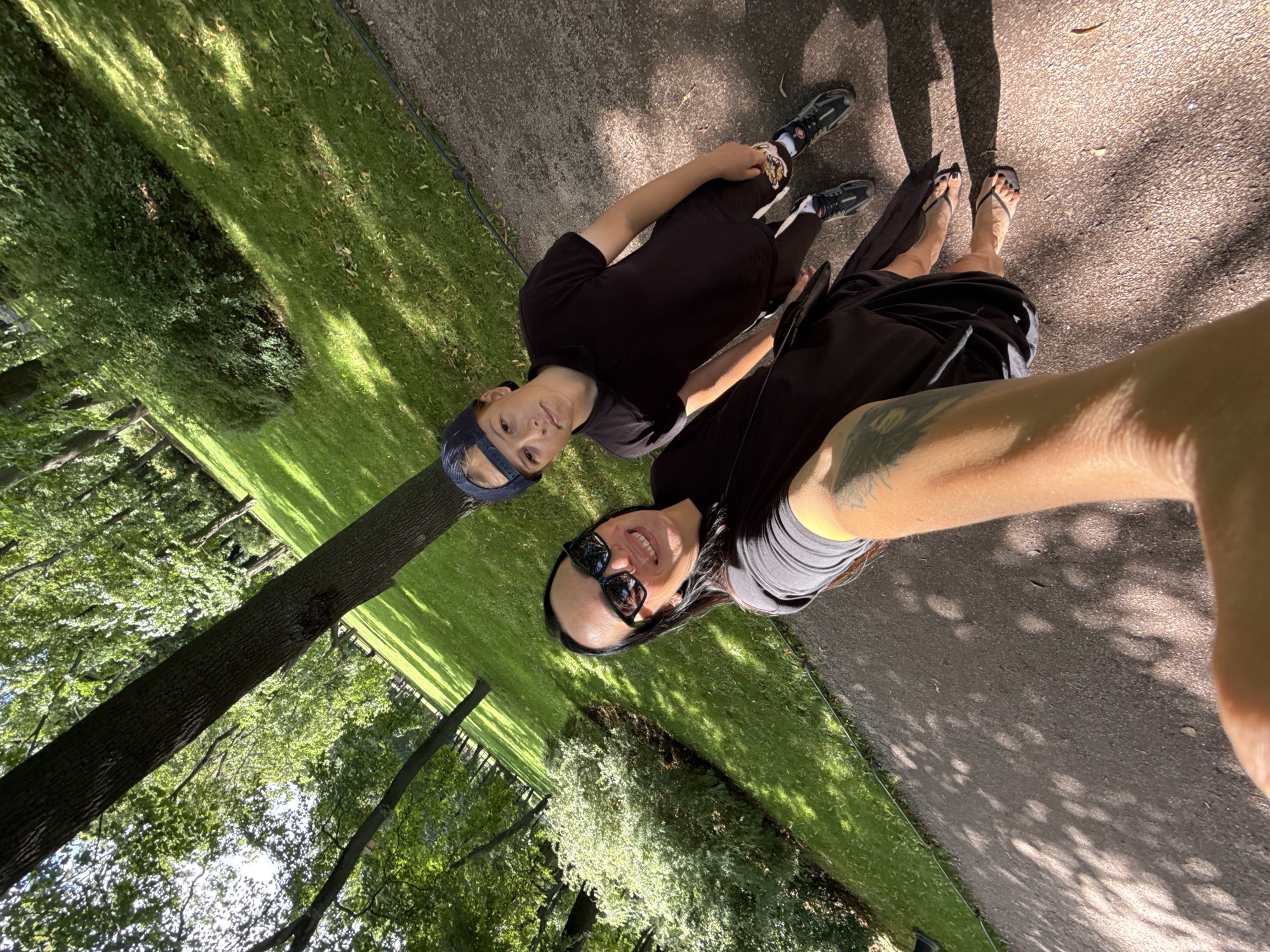 A selfie taken in a park shows a woman and a boy standing on a paved path. The woman, in the foreground, smiles at the camera. She is wearing sunglasses and a dark dress. The boy, standing behind her and to her right, looks towards the camera. He is wearing a black polo shirt, dark shorts with white stripes, a blue baseball cap, and sneakers. The park features a grassy slope covered in dappled sunlight and trees. The image is taken from a high angle, looking down at the subjects. The location is the Park of the 300th Anniversary of Saint Petersburg, Russia.