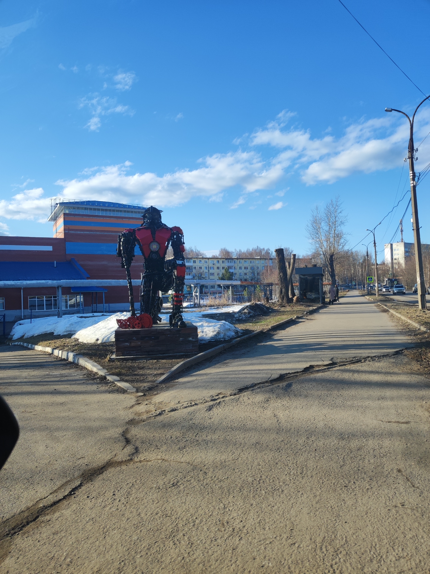 The image depicts a street scene in Votkinsk, Russia. On the left side of the road, a large metal sculpture of a robot-like figure stands on a wooden base. The figure is primarily dark metal with red accents on the chest and shoulders, holding what appears to be a large, decorative hammer. Patches of melting snow are visible around the base of the sculpture.
Behind the sculpture is a large building with a blue and orange striped facade. The right side of the image shows a road with a sidewalk, bare trees, and utility poles lining the street. A bus stop shelter is visible further down the road. Vehicles are seen driving in the distance.
The sky is blue with scattered clouds, suggesting a sunny day. The road surface is cracked and shows signs of wear. The overall scene suggests a post-winter, early spring setting.