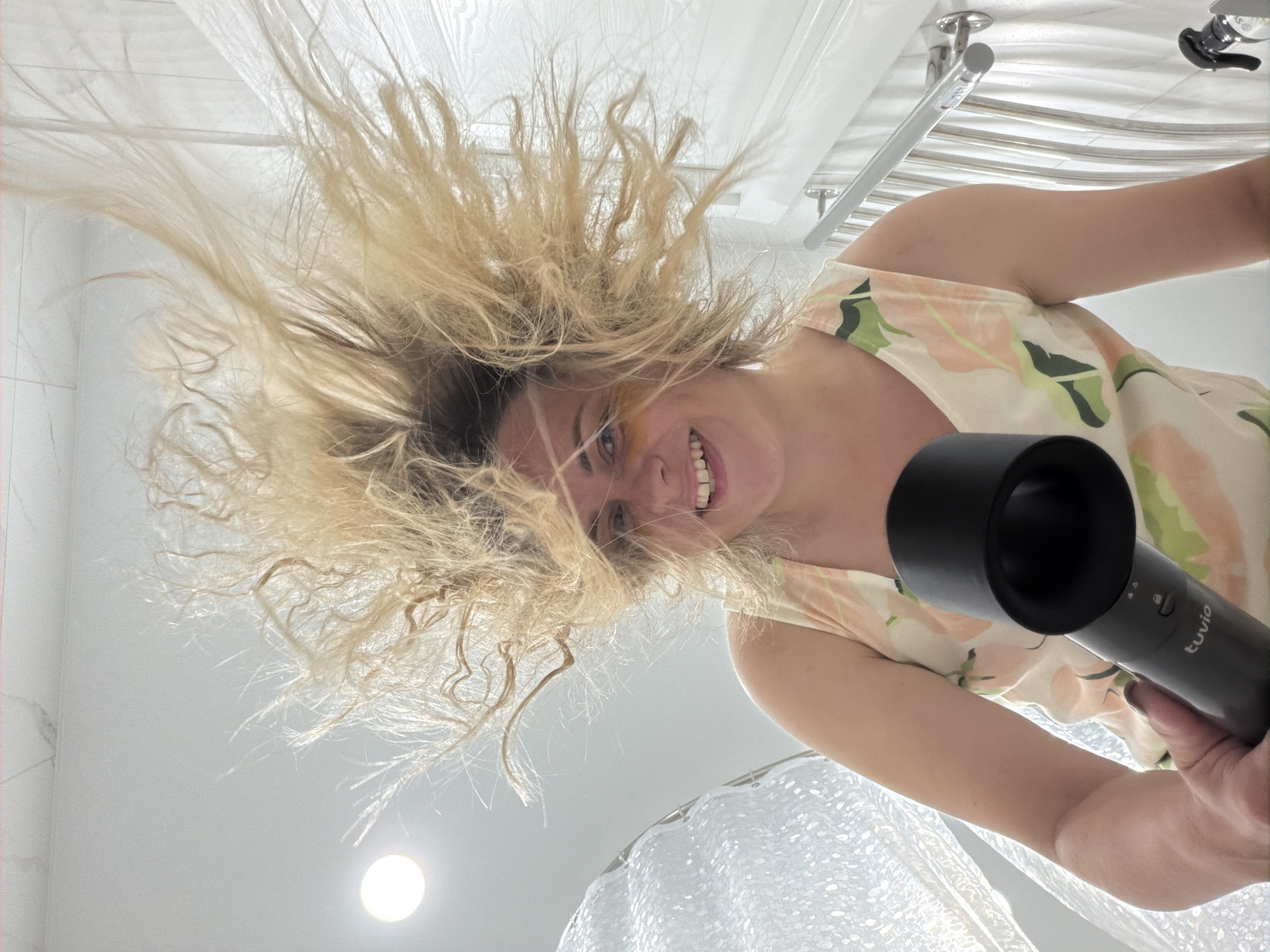 The image shows a woman with her blonde hair blown outwards by a hairdryer. She is smiling and holding the hairdryer in her right hand. She is wearing a sleeveless dress with a floral pattern. The background appears to be a bathroom with white tiled walls and a metal towel rack. There is a circular light fixture on the ceiling. The overall impression is of a playful moment captured during hair drying. The location in the city of Votkinsk, Russia, is not discernible from the image.