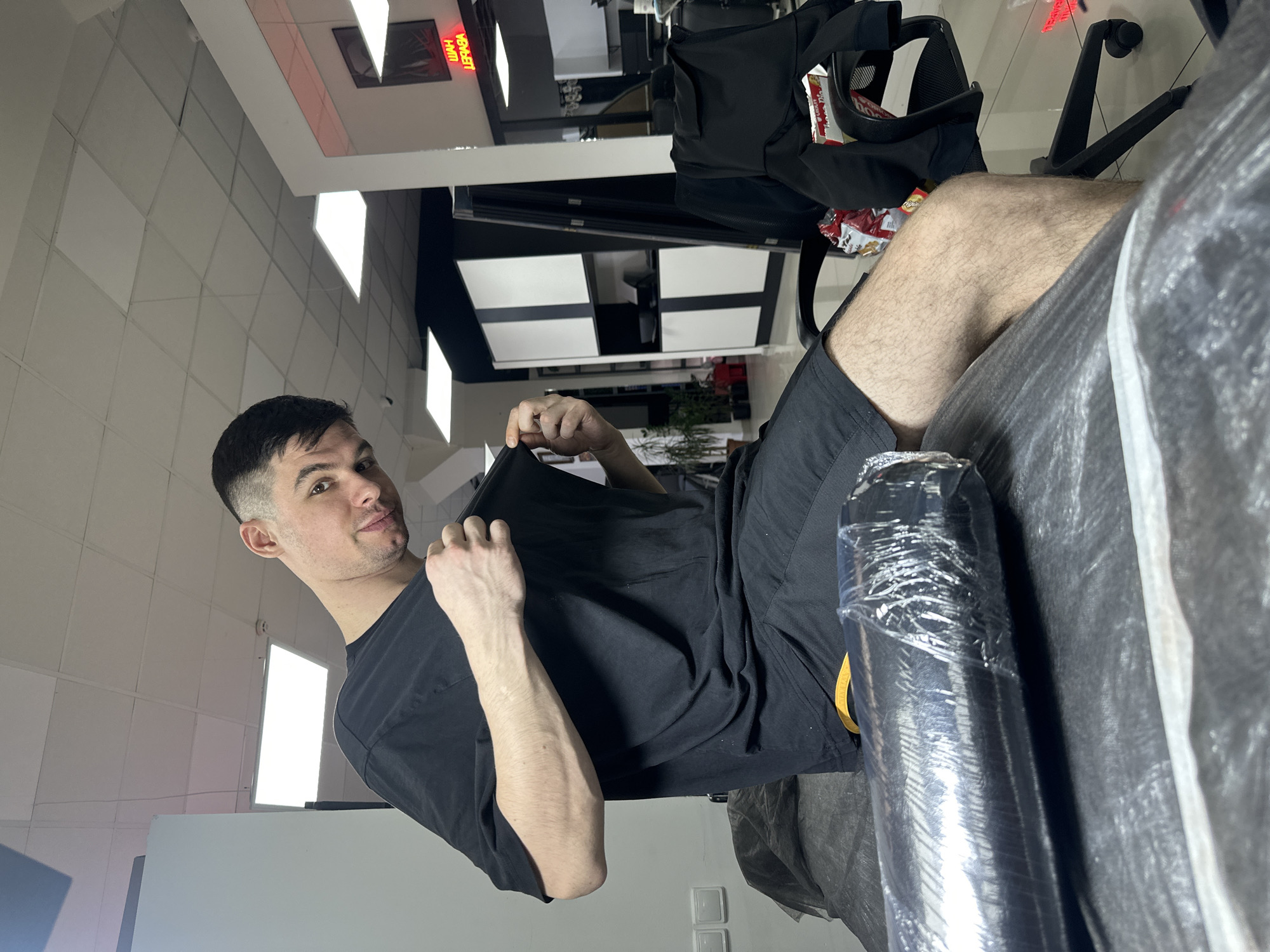 A man is sitting on a tattoo bed in what appears to be a tattoo parlor in Rostov-on-Don, Russia. He is wearing a black t-shirt and black shorts, with part of his bare leg visible. He holds his t-shirt up in front of him. A roll of wrapped plastic lies on the bed beside him. An office chair, with a black bag and snack items, is visible in the background. The parlor has tiled floors and ceiling panels with inset lighting. A sign with the word "работаем" (we are working) is visible in Russian. A black and white wall cabinet is present in the background.