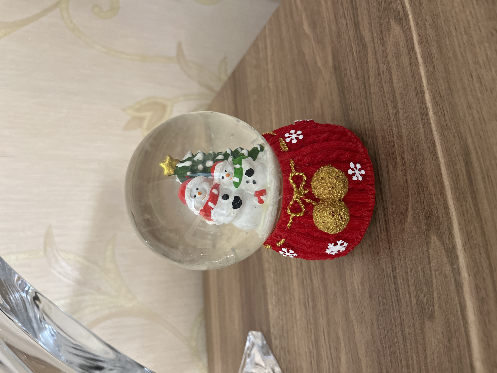 A Christmas snow globe sits on a wooden surface. The snow globe has a clear glass sphere containing two snowmen, a Christmas tree, and a gold star. The base of the snow globe is red with white snowflake decorations and two gold pom-poms. The background is a wall with a patterned wallpaper. The scene is illuminated by ambient light.