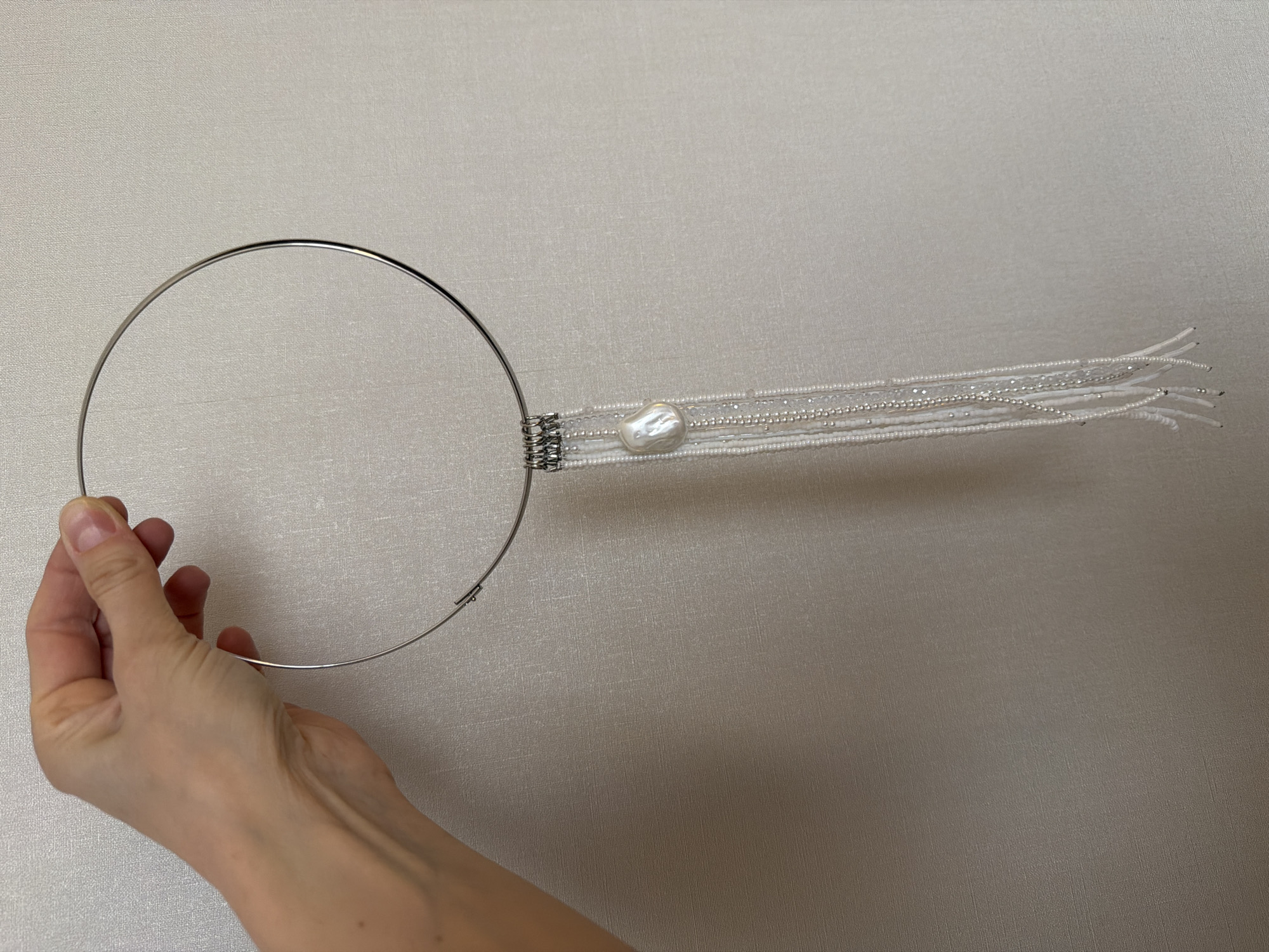 A hand is holding a silver metal circular choker necklace with a white pearl and beaded strand attached. The background is a light beige textured wall. The location and information about Moscow, Russia, cannot be determined from the image.