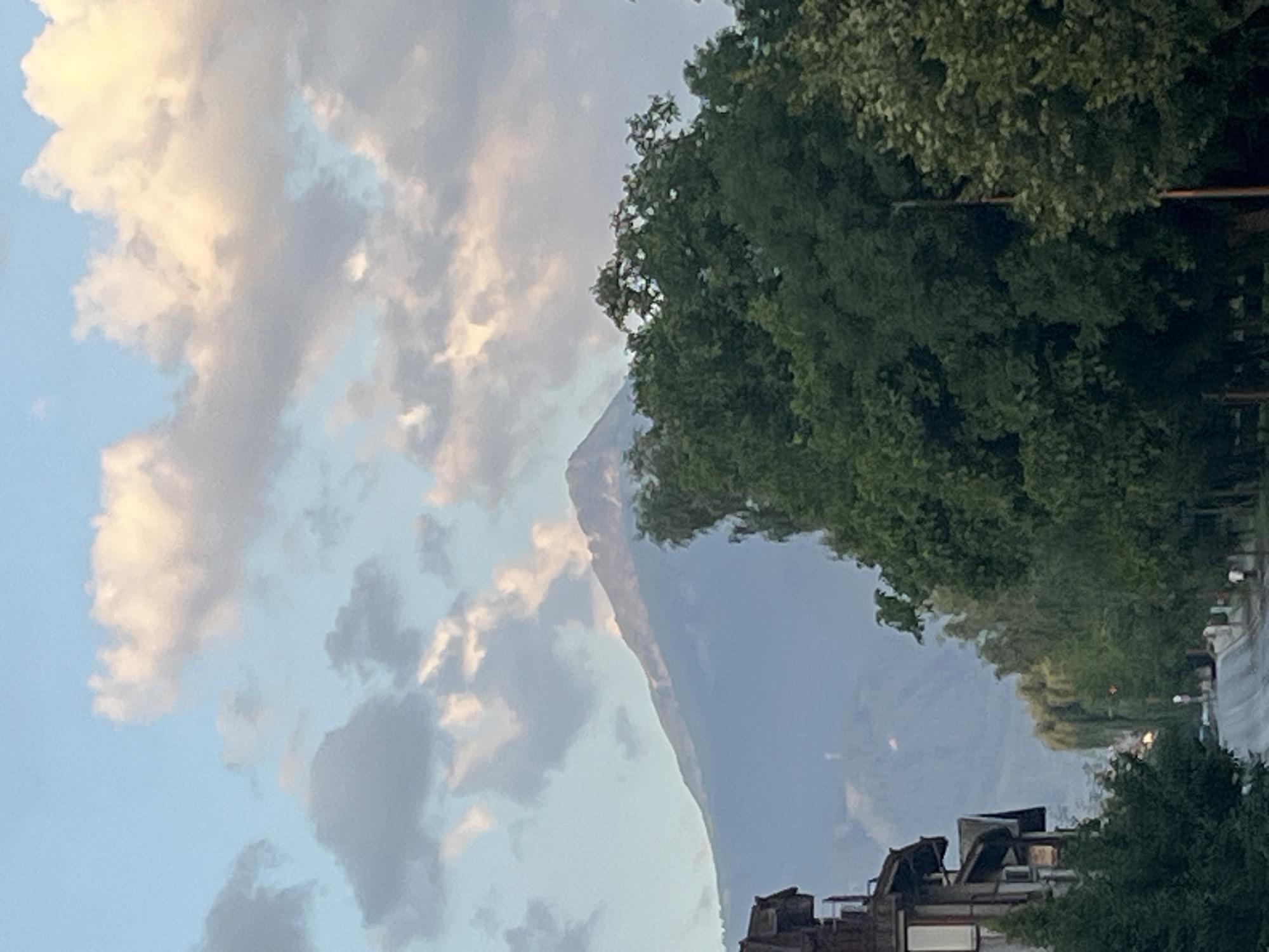The image depicts a street scene in Bansko, Bulgaria, with a towering mountain range in the background. The sky above is a soft blue, punctuated by fluffy white and gray clouds, with hints of warm light suggesting either sunrise or sunset. In the foreground, a multi-story building with a dark, pitched roof and light-colored walls stands to the left, bordering a tree-lined street. The street itself appears to be paved and curves gently away from the viewer, with a few indistinct figures visible walking along its edge. Lush green trees dominate the midground, with a dense canopy of foliage partially obscuring the mountain view. The mountain, hazy and majestic, rises sharply in the distance, its peak catching the light.