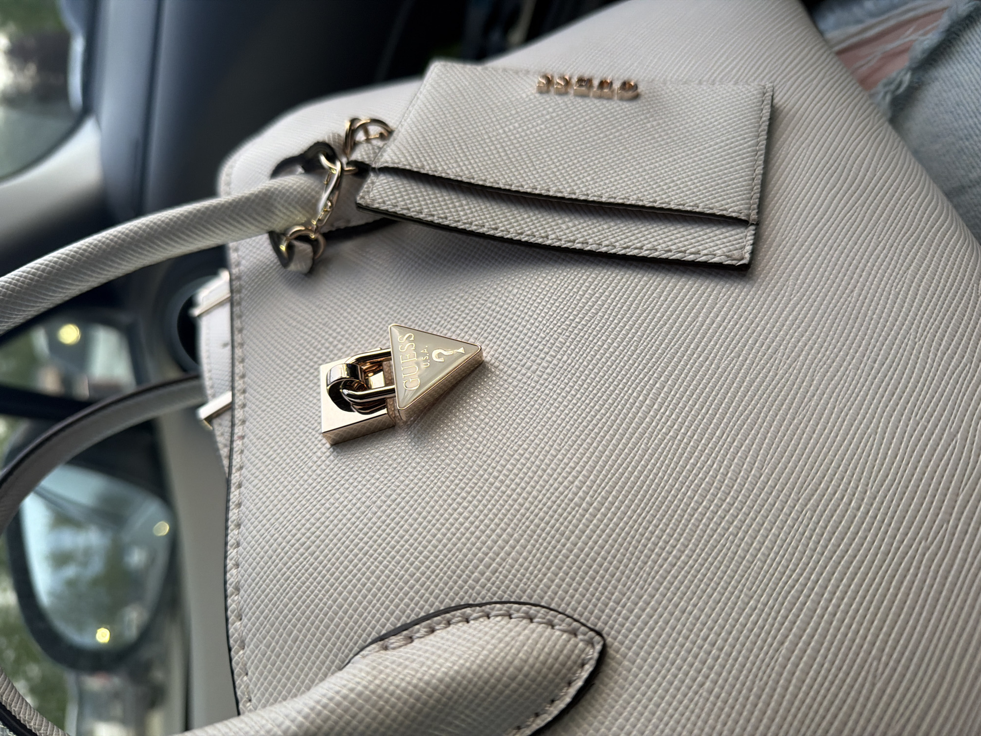 The image shows a close-up of a Guess handbag, light gray in color, with a textured surface. A gold-colored Guess logo and padlock are visible. The handbag is likely inside a car, given the blurred view of a window and the car's interior in the background. There is no information to determine if the image was taken in Krasnoyarsk, Russia.