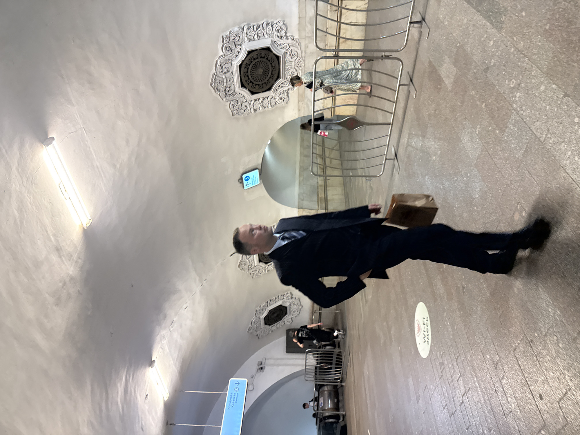 The image depicts a man in a dark suit walking through what appears to be a subway station in Moscow, Russia. He is holding a brown paper bag in his right hand and has his left hand in his pocket. The man is in the foreground, slightly out of focus. In the background, other people are visible, including a woman in a patterned dress and a person wearing a backpack. There are also subway signs, metal barriers, and architectural elements characteristic of a metro station, such as curved ceilings and ornate decorative panels. A circular marking on the floor indicates a "Wi-Fi" zone. The lighting suggests an indoor environment, likely underground.