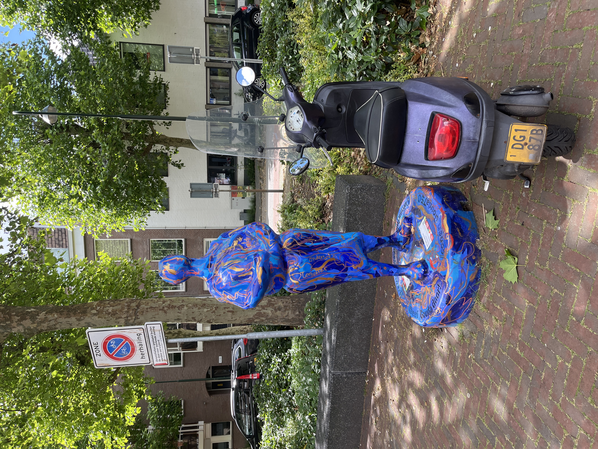 In an outdoor urban setting, likely a park or sidewalk, a vibrantly colored sculpture of a standing figure, painted in swirling patterns of blue, orange, and gold, is positioned next to a dark purple motor scooter. The sculpture is placed on a circular base, which also displays the same artistic patterns. The scooter is facing to the right, with its license plate, DG1 87B, clearly visible. Behind the sculpture and scooter, a dense arrangement of green bushes and trees provides a natural backdrop, interspersed with urban architecture including apartment buildings and a street lamp. A traffic sign with a red circle and a diagonal bar, indicating a zone where parking is prohibited for bicycles and mopeds, is visible to the left of the sculpture. The ground is paved with reddish-brown brickwork laid in a herringbone pattern. Sunlight filters through the trees, casting dappled shadows on the ground.