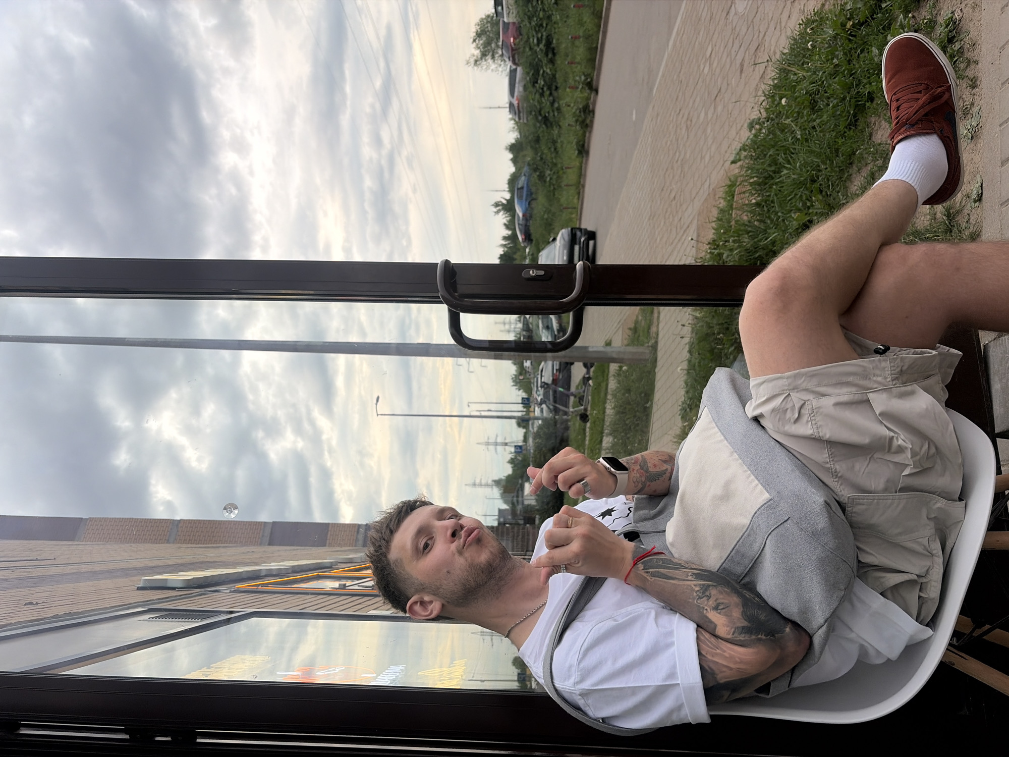 A young adult male is seated on a white chair, leaning back against the glass facade of a building, likely a commercial establishment identified by the reflected "СТОЛОВАЯ" (Stolovaya - Canteen/Cafeteria) sign. He is situated on a brick-patterned sidewalk in Saint Petersburg, Russia. The man has short brown hair, stubble, and is wearing a white short-sleeved t-shirt, light khaki cargo shorts, white ankle socks, and reddish-brown suede sneakers. He displays visible black and grey tattoos on both forearms, depicting what appear to be classical figures or portraits. Accessories include a silver chain necklace, a gold wedding band on his left ring finger, a silver ring on his right index finger, a white smartwatch on his left wrist, and a red string bracelet on the same wrist. A grey and beige two-tone tote bag with a strap over his left shoulder rests on his lap. He is looking directly at the camera with pouting lips and holds both hands raised, forming a "finger heart" gesture with his thumbs and index fingers.
Beyond the man and the building's glass, a cloudy, overcast sky dominates the upper frame. The background reveals an urban or suburban landscape: a paved road with a visible dark lamppost on the right, and a grassy embankment separating the road from what appears to be a parking area or industrial zone. Multiple vehicles (including a blue SUV and other cars) are visible on and off the road. Several power line towers and overhead lines traverse the horizon in the middle distance. A dark brown door handle set is visible on the right side of the glass panel near the man.