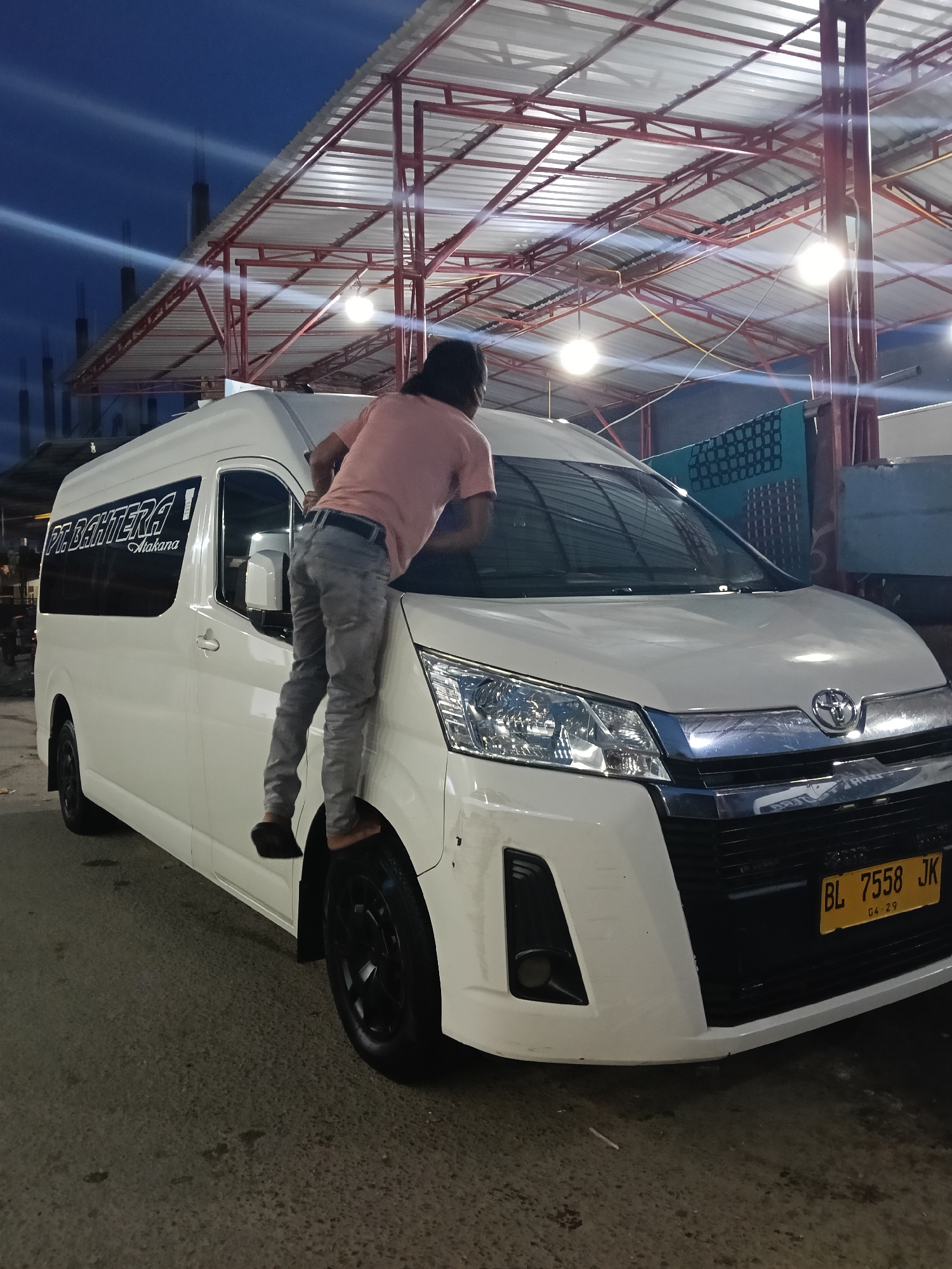 A person is standing on the tire of a white Toyota Hiace van, cleaning its windshield under a metal structure with lights. The van has black rims, tinted windows, and a license plate that reads "BL 7558 JK." The sign on the van's window reads "PT. BAHRETERA Atakana." The scene appears to be in an urban setting at dusk or night. Based on the license plate, the location is Lhokseumawe, Indonesia. The surroundings show construction in the background. The person is wearing a pink shirt and light-colored pants.
