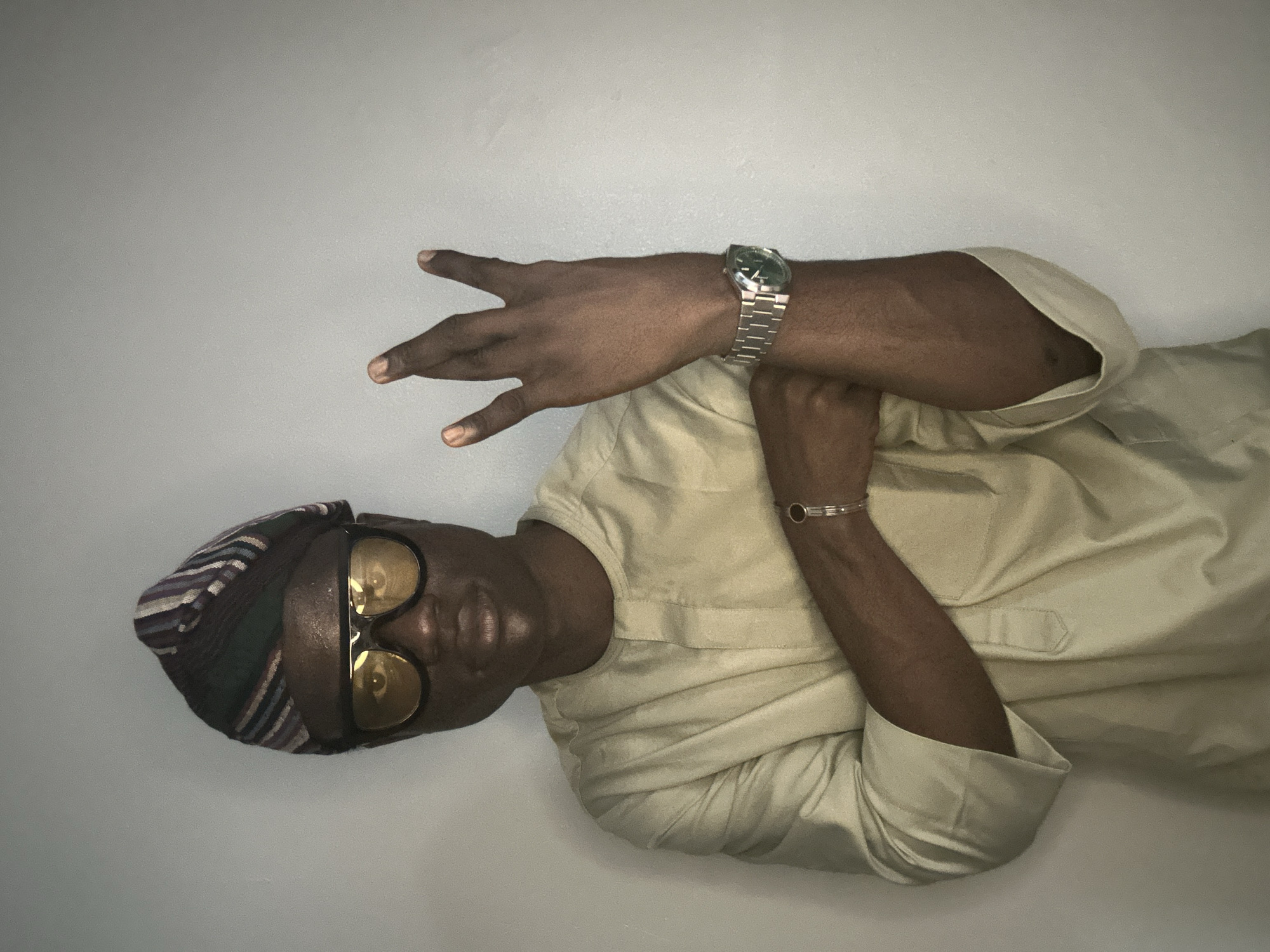 A person is standing in front of a gray wall, wearing a traditional Nigerian outfit and sunglasses. The person has on a light-colored, long-sleeved shirt with a Mandarin collar. On their head, they are wearing a striped cap. They are also wearing large, dark-rimmed sunglasses with yellow lenses. Their left arm is crossed over their chest, and they are wearing a silver watch on their left wrist and a silver bracelet on their right wrist. The person is holding up their right hand, with three fingers extended. The lighting in the image is dim, casting shadows on the person's face and arms. The background is a plain, textured gray wall. The scene does not contain any elements that place it specifically in the city of Badagry, Nigeria.