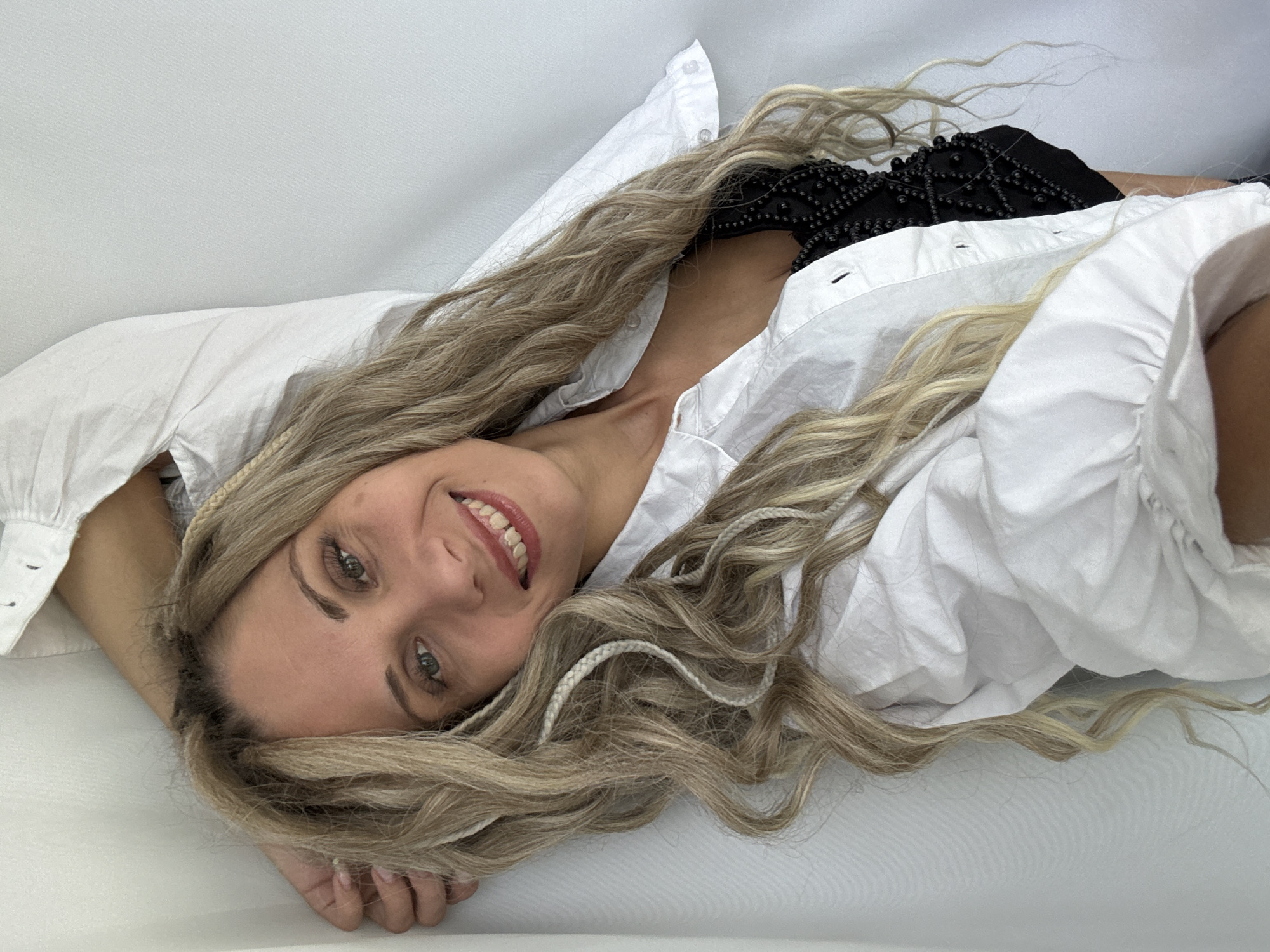 A woman lies on a white surface. She is wearing a white shirt, and her hair is long and blonde with braids. Part of a black, beaded garment is also visible.