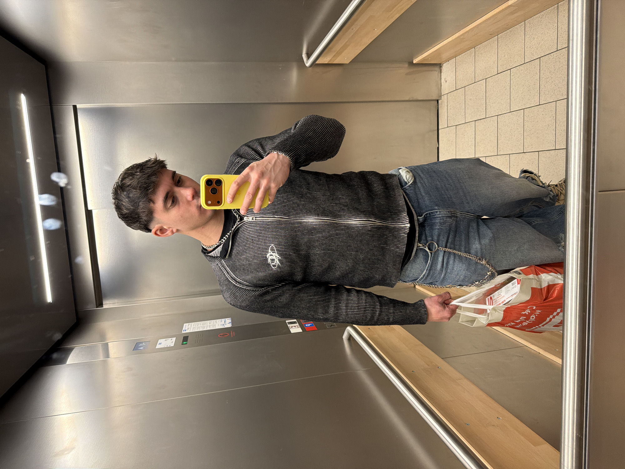 A male subject is depicted in an elevator, taking a mirror selfie. He has short, dark hair with faded sides. He wears a dark, vertically ribbed zip-up jacket with a white logo on the left chest and a silver chain around his neck. His lower attire includes distressed denim jeans with a silver wallet chain and partially visible light-colored sneakers. In his right hand, he holds a bright yellow smartphone for the selfie. His left hand carries a white and orange shopping bag bearing the Cyrillic text "Фантастико". The elevator walls are reflective silver metallic panels. A control panel on the left wall features illuminated buttons, with the '9' button glowing red. A horizontal LED light strip illuminates the ceiling. A silver handrail with a light wood base spans the lower section of the wall. Light-colored tiles form the floor. Mirror smudges are visible. The "Фантастико" shopping bag indicates the scene is in an elevator within or connected to a Fantastico supermarket or an associated commercial/residential building in Sofia, Bulgaria.