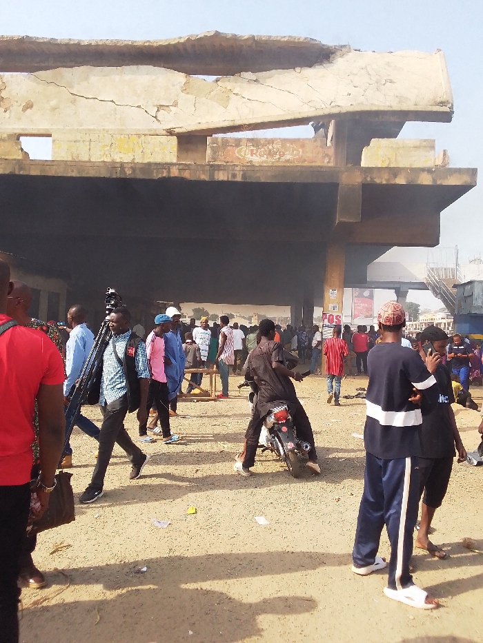 The image shows a scene in Jos, Nigeria, at the coordinates 9.921 latitude and 8.89 longitude. There are approximately 30 people standing in a large, open space beneath a partially collapsed concrete structure. Some of the people appear to be observing the collapsed section of the structure while others stand in casual conversation. A man is sitting on a black motorcycle with a second person behind him. There is a camera on a tripod in the foreground of the image. The ground is dusty and there are signs of debris on the ground. The sky is clear and blue. The collapsed concrete section appears to have been part of an elevated roadway or walkway. There is a second concrete structure visible in the background that appears to be a bridge or overpass. The scene is bright and sunny, with shadows cast by the people and structures. A large portion of the concrete structure appears to have been charred, indicating possible damage caused by a fire. The scene is likely chaotic and there is a sense of urgency and concern among the people. There is no sign of emergency responders or authorities in the image.