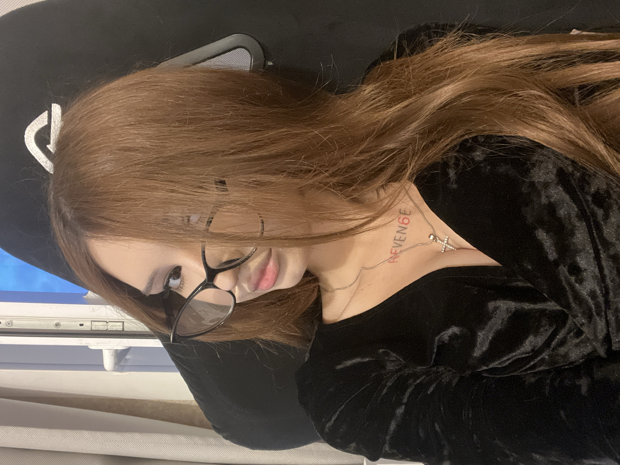 A young woman with long, auburn hair and glasses is looking into the camera. She is wearing a black velvet dress and a silver necklace with a cross pendant. She has a tattoo on her chest that says "REVENGE". The background is dark and appears to be an interior setting.