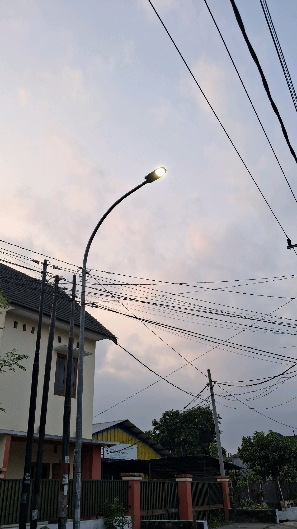 The image depicts a street scene in Banguntapan, Indonesia, likely during the early morning or late evening given the illuminated streetlamp and the soft sky colors. In the foreground, a tall, curved streetlamp is lit, its light casting a glow. A dense network of overhead power lines crisscrosses the sky, supported by utility poles. To the left, a section of a two-story house is visible, featuring a dark tiled roof, cream-colored walls, and windows with dark frames. In front of the house, several dark, vertical poles stand, possibly part of a fence or utility infrastructure. Further in the background, a building with a yellow facade and a corrugated metal roof is visible, along with more trees and other structures suggesting a residential or mixed-use area. The overall impression is a typical urban or suburban streetscape with abundant electrical infrastructure.