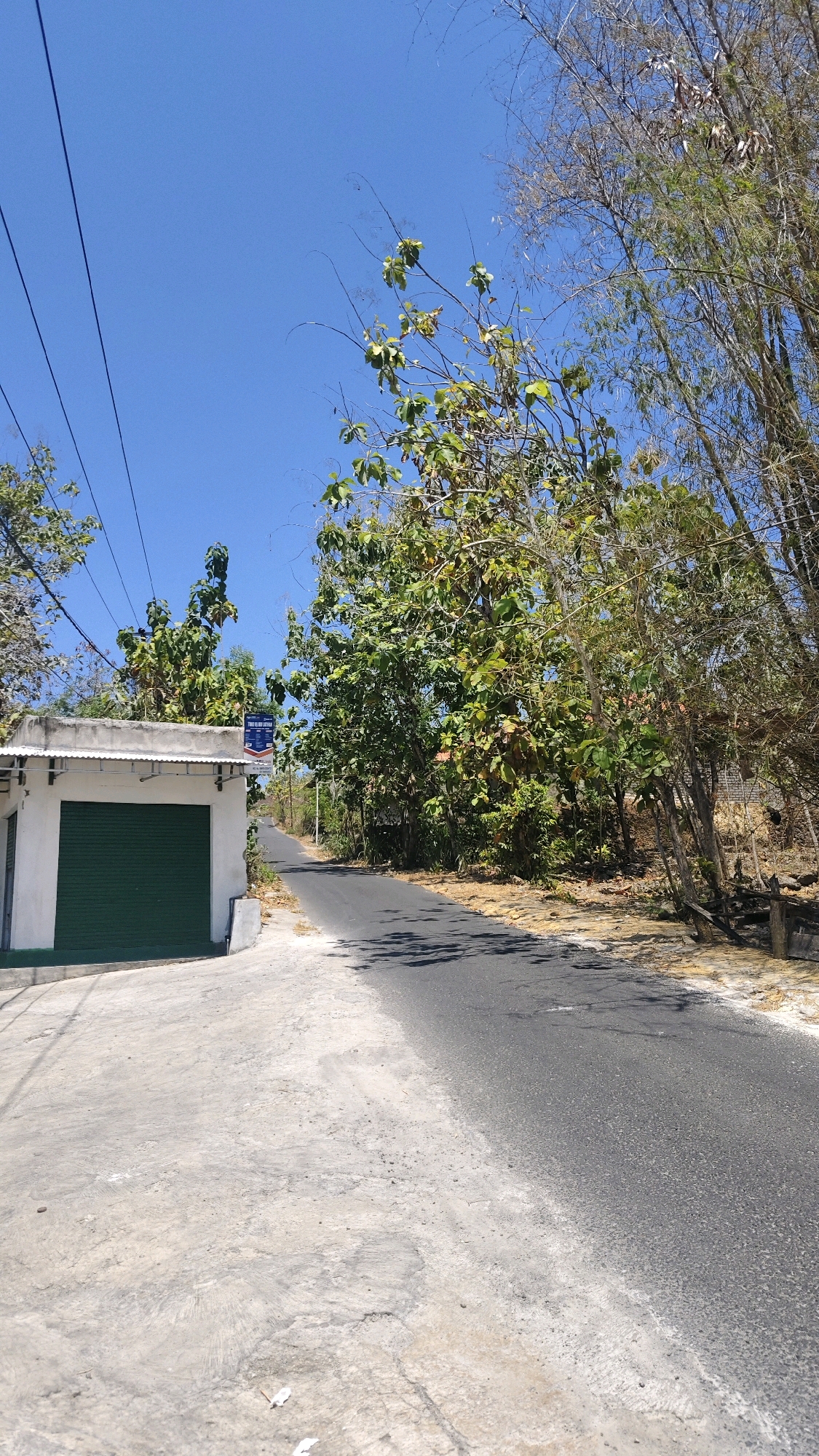 The image depicts a street scene in Tellok, Indonesia, on a clear, sunny day. On the left side of the road, a small, white, single-story building with a green roll-up door is visible. Above the building, power lines stretch across the frame against a bright blue sky. The road itself is paved and curves to the right, disappearing into the distance. The right side of the road is lined with dense foliage, including trees with green leaves and bamboo stalks. The ground is a mix of concrete and dirt. A sign with blue and white lettering is mounted on the building. No people are present in the image.