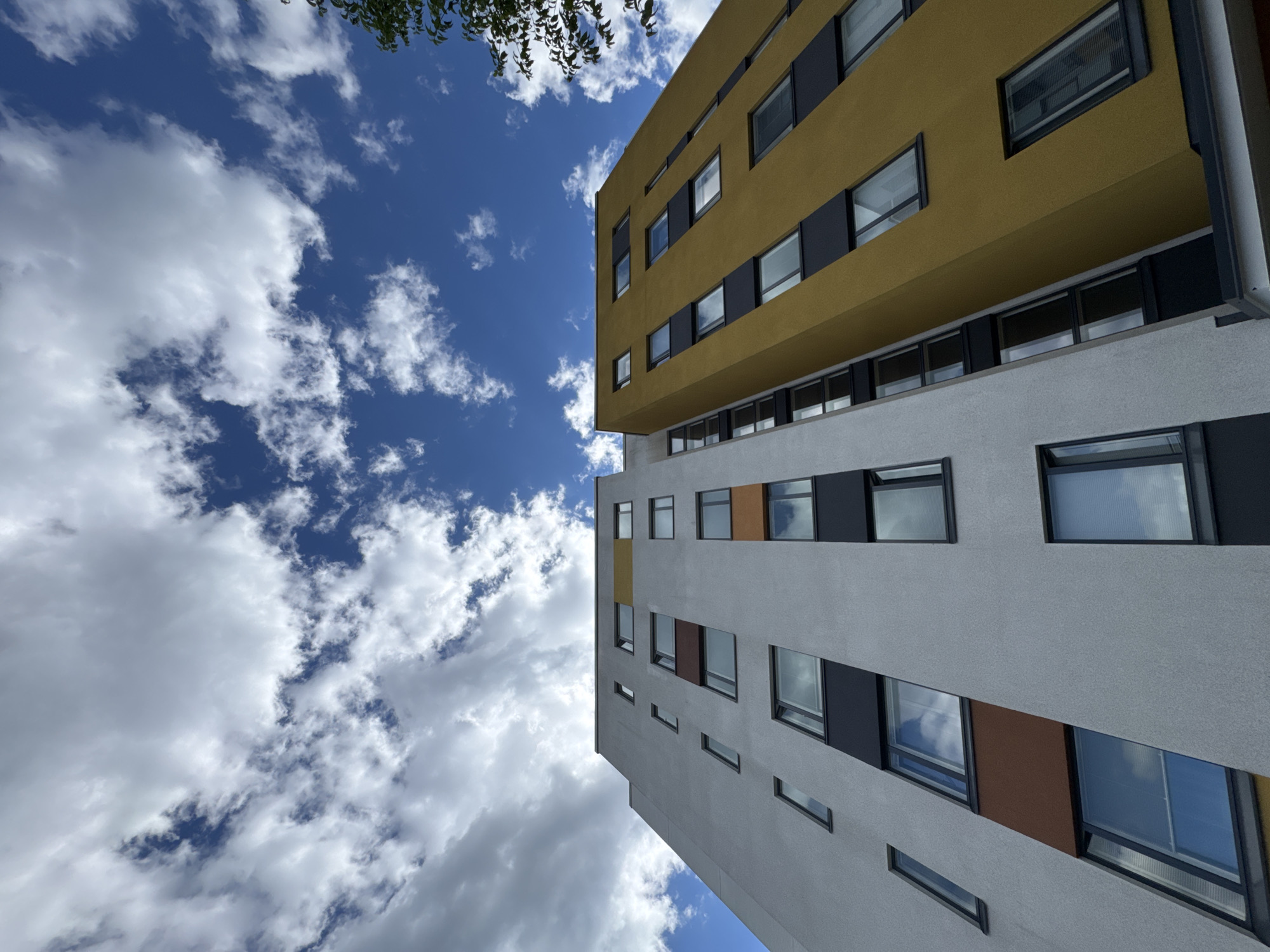 This media file is a low-angle, wide shot of a modern building against a blue sky with white clouds. The building has multiple stories and features a mix of yellow and grey exterior panels, with numerous windows. Some windows are large and rectangular, while others are smaller and square. Dark grey or black frames surround the windows. The upper section of the building is a vibrant mustard yellow, contrasting with the lighter grey of the lower section. There are also accents of dark grey and a few panels of brown or rust color visible on the grey section. The building appears to be an apartment complex or an office building due to its uniform window structure. The sun is high in the sky, suggesting it is daytime, and the presence of fluffy white clouds indicates fair weather. The perspective is looking up from the ground, emphasizing the height and architectural design of the building. There is a hint of green foliage from a tree in the upper left corner of the frame, suggesting an outdoor setting with some vegetation. No people or specific activities are depicted, only the architectural structure and the natural elements. No visible text is present in the image.