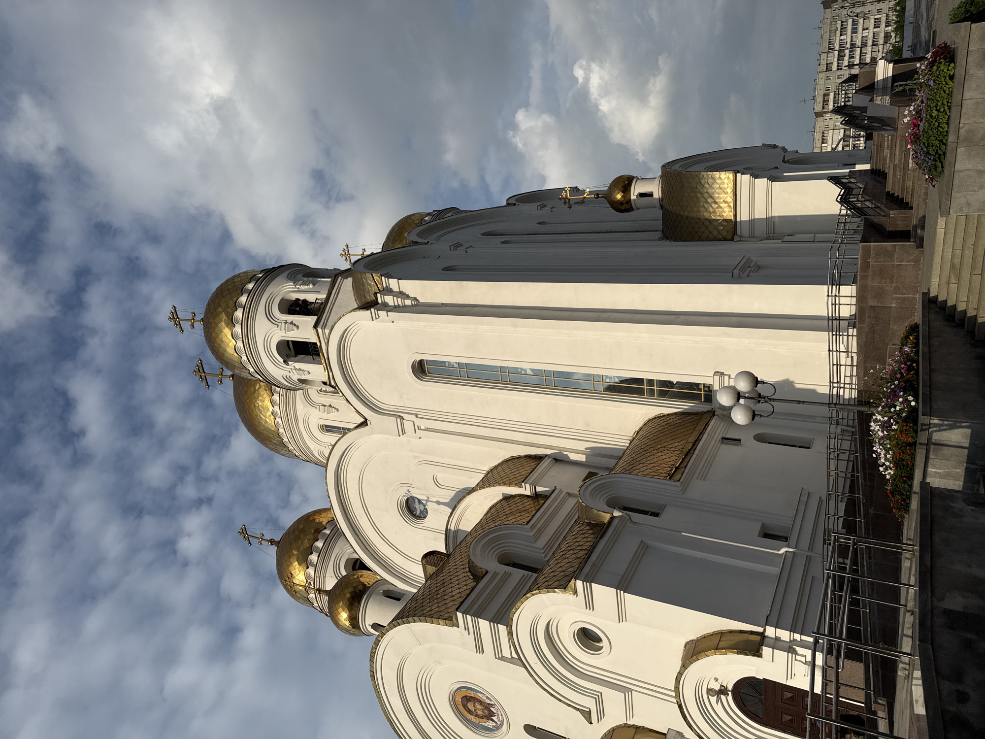 A low-angle shot captures a grand, white Orthodox church with numerous golden domes topped with crosses. The church features arched windows and intricate architectural details. A prominent fresco of Jesus Christ is visible on the left side of the building, encased in a circular frame. In the foreground, steps lead up to the entrance, flanked by metal railings and flowerbeds. A street lamp with multiple globes stands to the right of the steps. The sky is filled with dramatic, partly cloudy skies, with sunlight casting shadows on the church. In the background, a modern, multi-story apartment building is visible, suggesting an urban setting. The overall impression is one of architectural grandeur and religious solemnity.