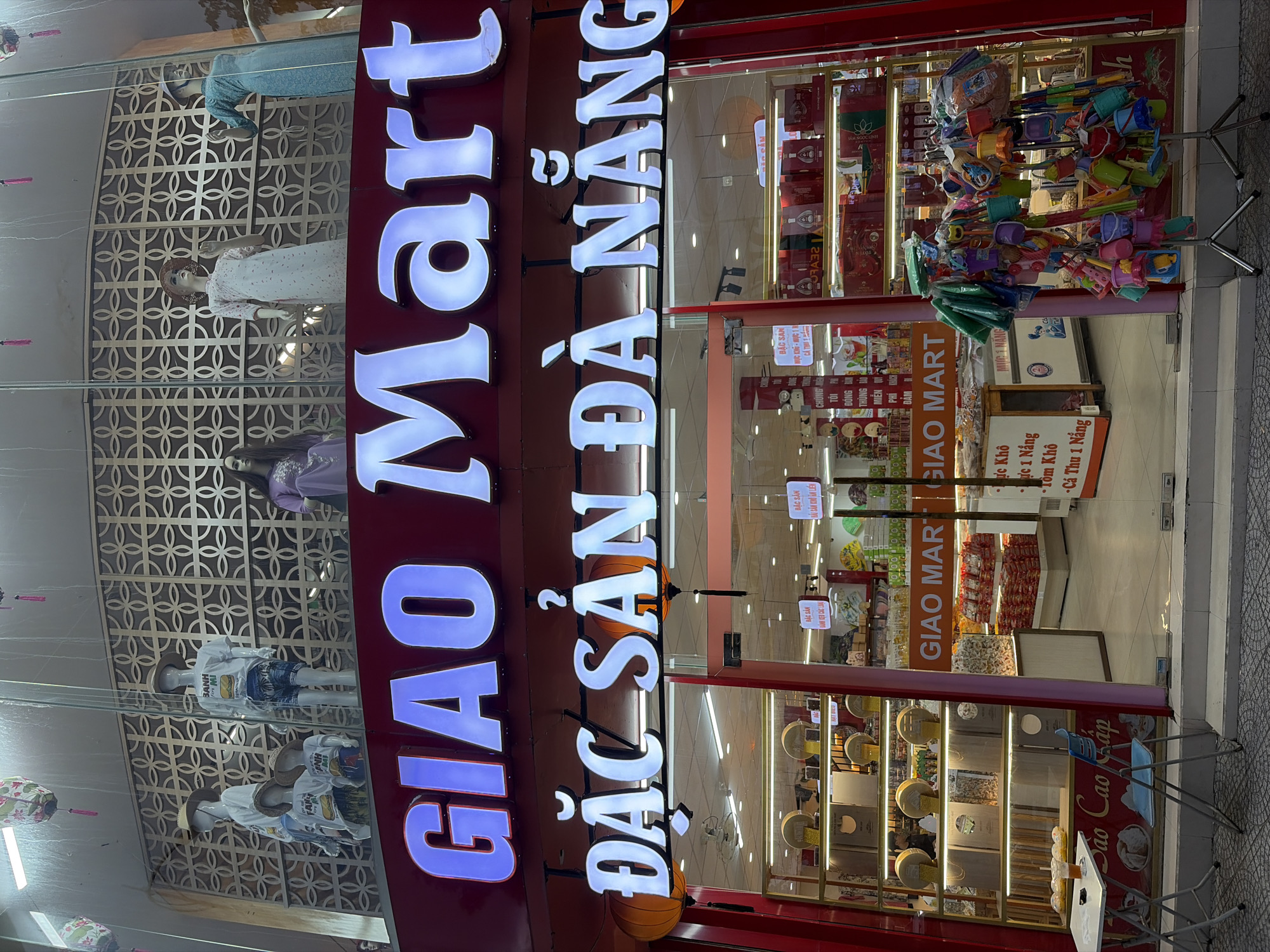 The image shows the storefront of a market named "Giao Mart" in the city of Đà Nẵng, Vietnam. The market's name is prominently displayed in large, illuminated letters on its facade. Below the main name, the text "ĐẶC SẢN ĐÀ NẴNG" is also visible, indicating that the market specializes in local specialties from Đà Nẵng.
The storefront features a large glass entrance, allowing a view into the interior. Inside, shelves are stocked with various products, including what appear to be packaged food items and other merchandise. Mannequins dressed in clothing are displayed in the upper windows. Two large orange lanterns are hung on either side of the "ĐẶC SẢN ĐÀ NẴNG" sign. Outside the entrance, a small table and chairs are set up, with a drink and some food items on the table. To the right of the entrance, a rack displays an assortment of colorful children's toys, such as buckets and sand molds. The overall scene depicts a retail environment in a Vietnamese city, likely in the evening or at night, given the illuminated signage.