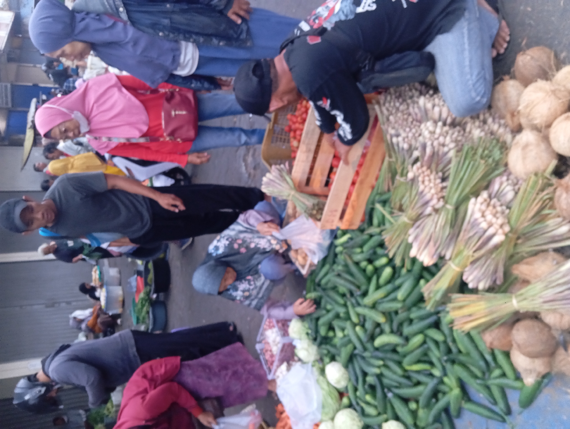 The image depicts an outdoor market scene, likely in Yogyakarta, Indonesia, characterized by stalls laden with fresh produce. In the foreground, a large pile of cucumbers is prominent, alongside bundles of lemongrass and several coconuts. To the right, a man wearing a black shirt and jeans, with a black baseball cap, is crouched down, appearing to sort or arrange tomatoes in a wooden crate. Behind him, a woman in a blue hijab and jacket stands, and another woman in a pink hijab and red top, also wearing a light-colored mask, is to her left, looking towards the produce. Further in the background, other individuals are visible, some engaged in market activities. The overall environment suggests a bustling marketplace with various vendors and shoppers.